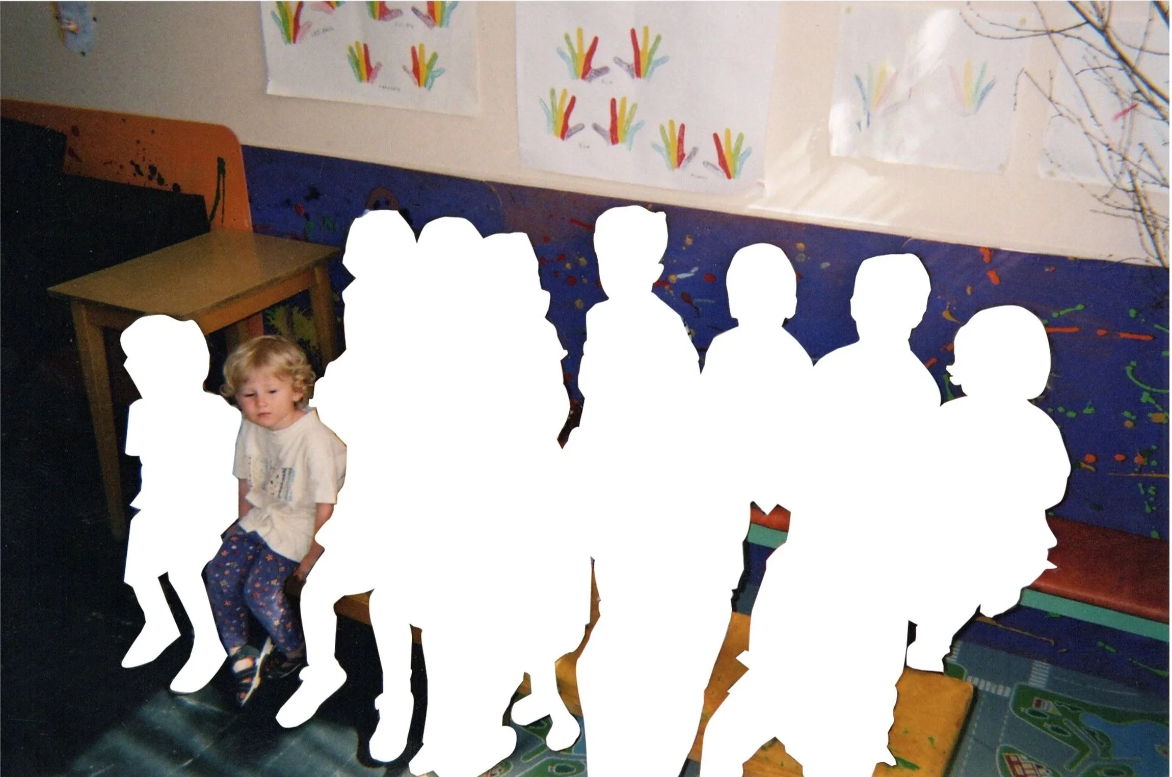 A young boy with curly blond hair sitting on a bench, with silhouettes of other children and adults behind him, some cut out or covered with white shapes, in a room with colorful children's artwork on the walls and a blue tablecloth with paint splatters on the table.