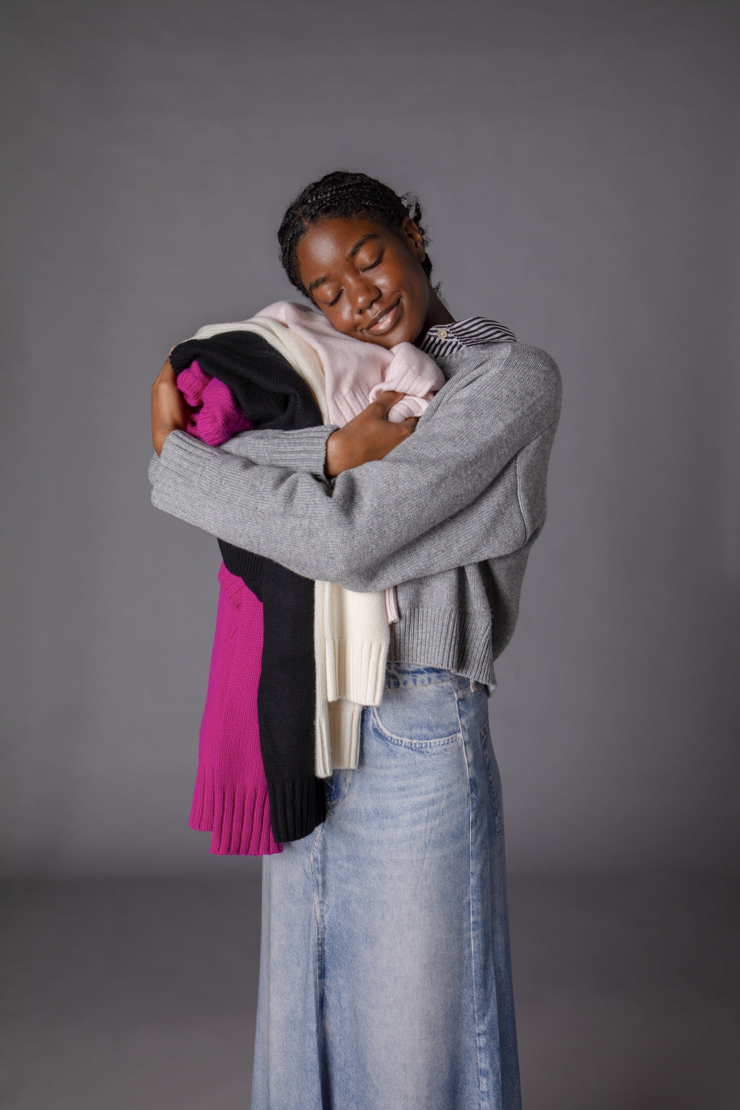 A woman with dark skin and braided hair, smiling with eyes closed, hugging a pile of folded clothes against a gray background.