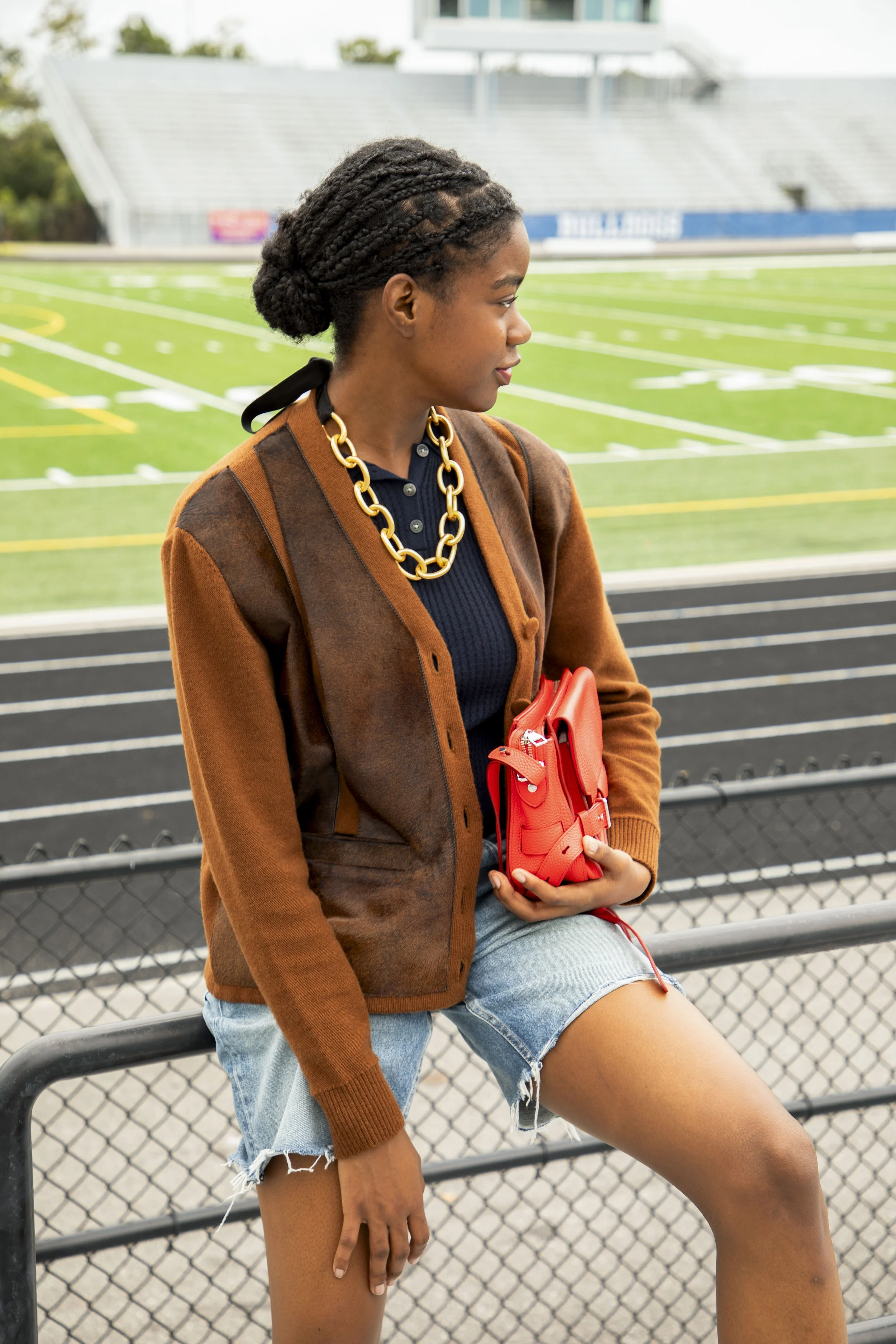 A young woman with braids, wearing a brown jacket, black top, and denim shorts, holding a red purse, standing by a fence at a football stadium.
