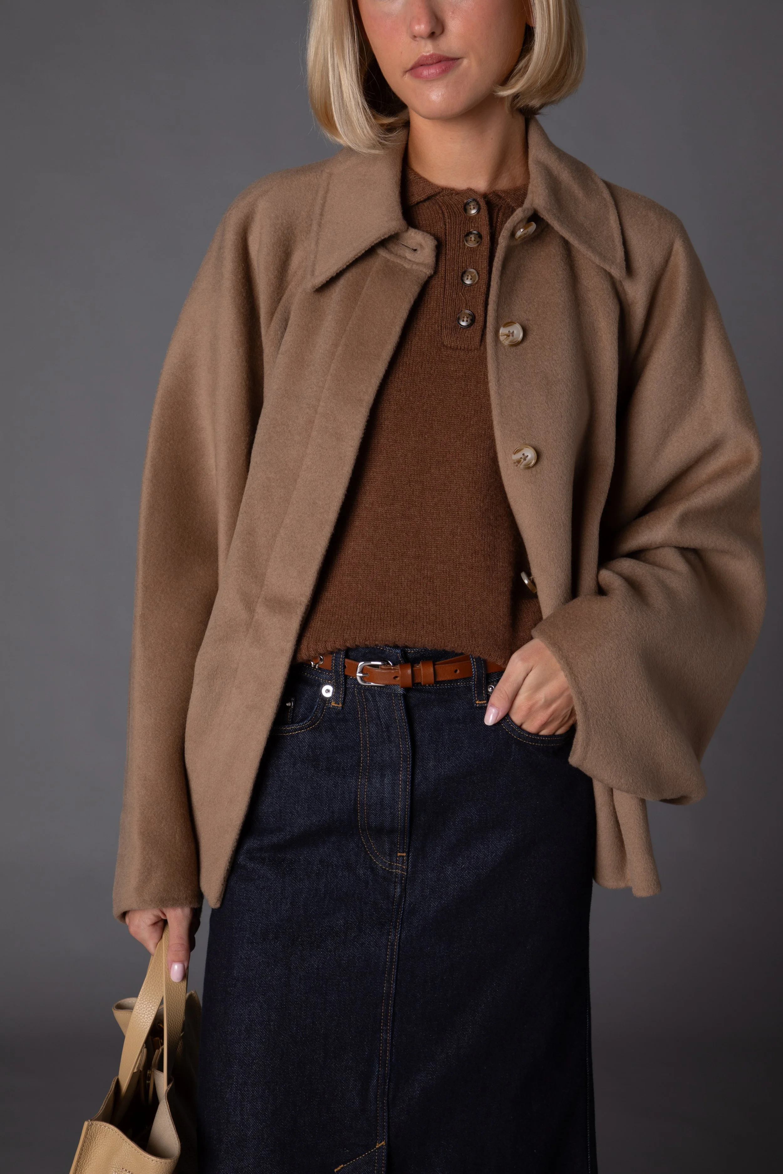 A woman wearing a brown coat over a brown top, with dark jeans and a brown belt, holding a beige handbag, standing against a gray background.