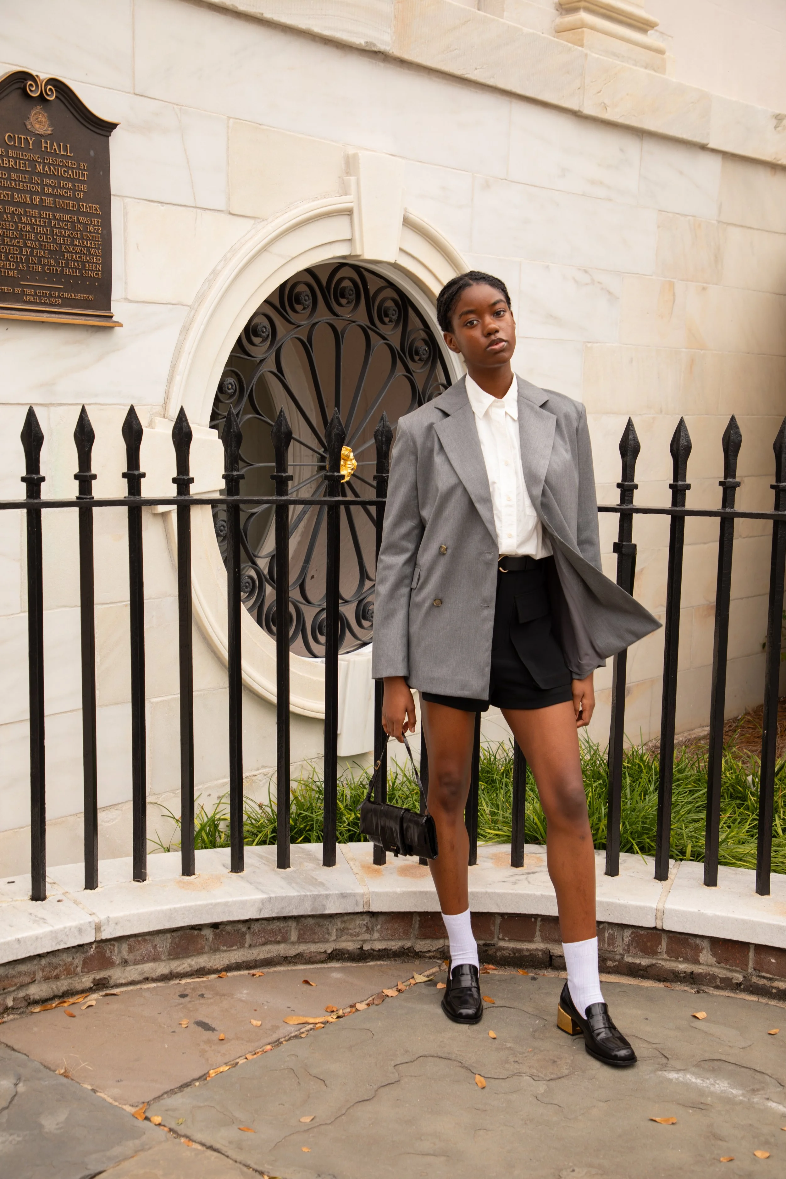 A young woman in a gray blazer, white shirt, black shorts, white socks, and black shoes standing in front of a historic building with a microphone and a black fence.