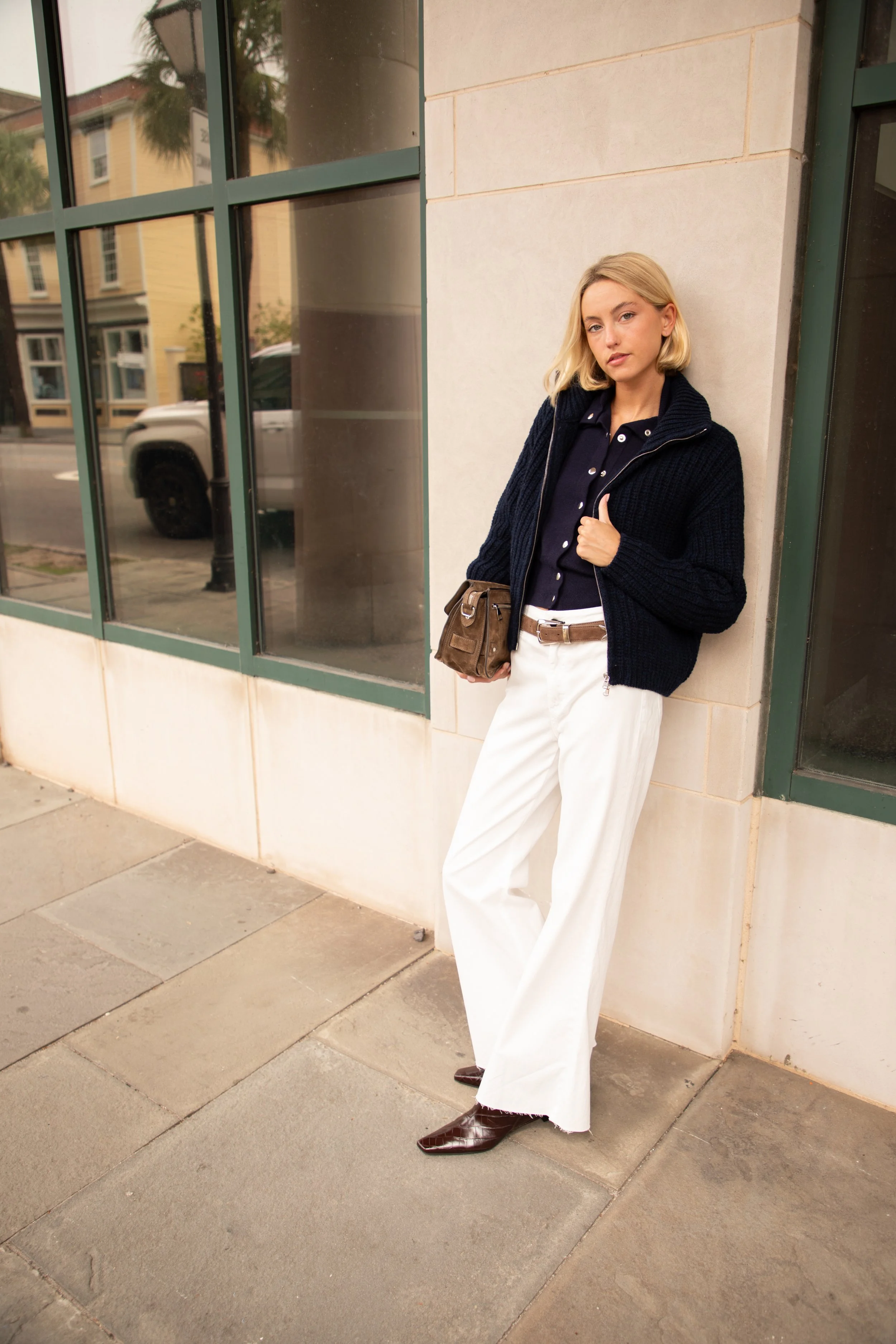 A woman with blonde hair wearing a navy blue jacket, black shirt, white wide-leg pants, and dark brown boots, standing against a beige wall near large glass windows with reflections of the street and parked cars.
