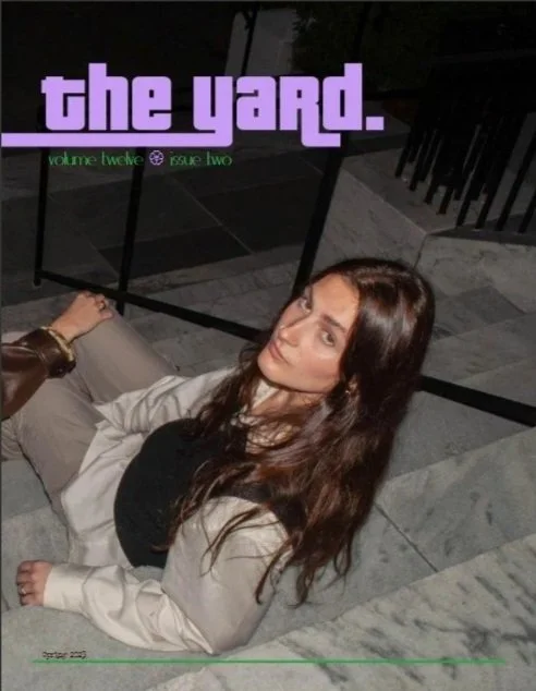 A woman with long brown hair sitting on stairs outdoors at night, looking at the camera. There is a magazine titled 'the yard.' with purple and green text in the background.
