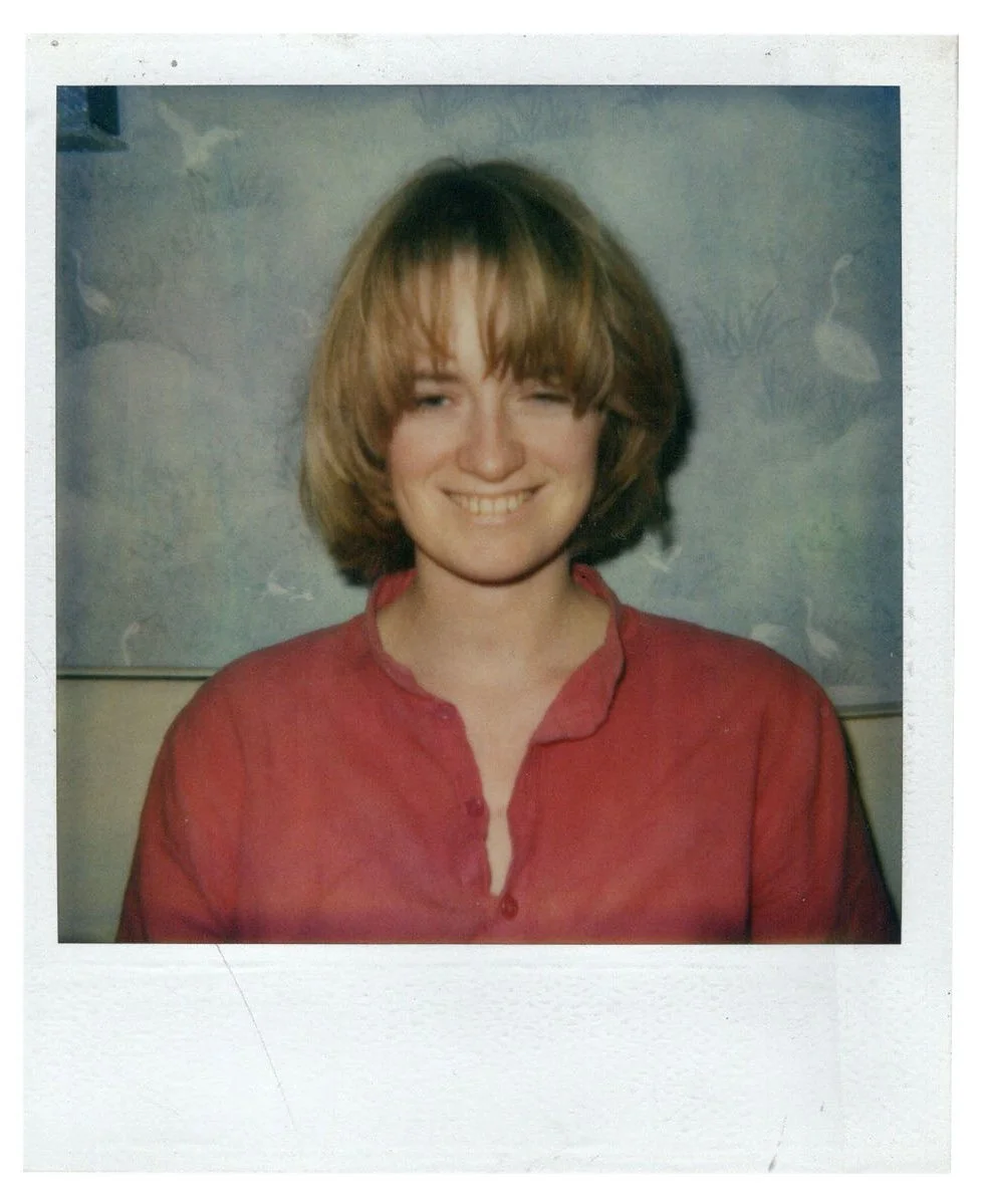 A woman with short, light brown hair smiling and winking, wearing a red button-up shirt, in front of a patterned background.