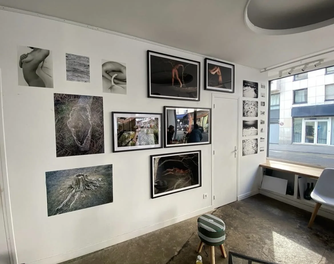 Interior view of an art gallery or studio with black and white photographs and artwork hung on white walls, a large window on the right, and a small stool on the floor.
