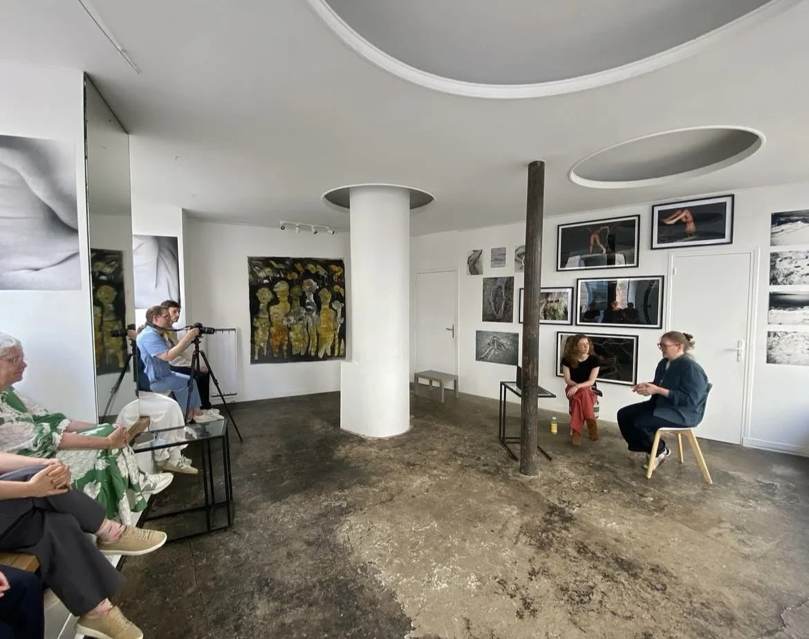 Two women sitting and talking in an art gallery during an interview, with paint photographs on the wall behind them, and a small audience and a photographer observing.