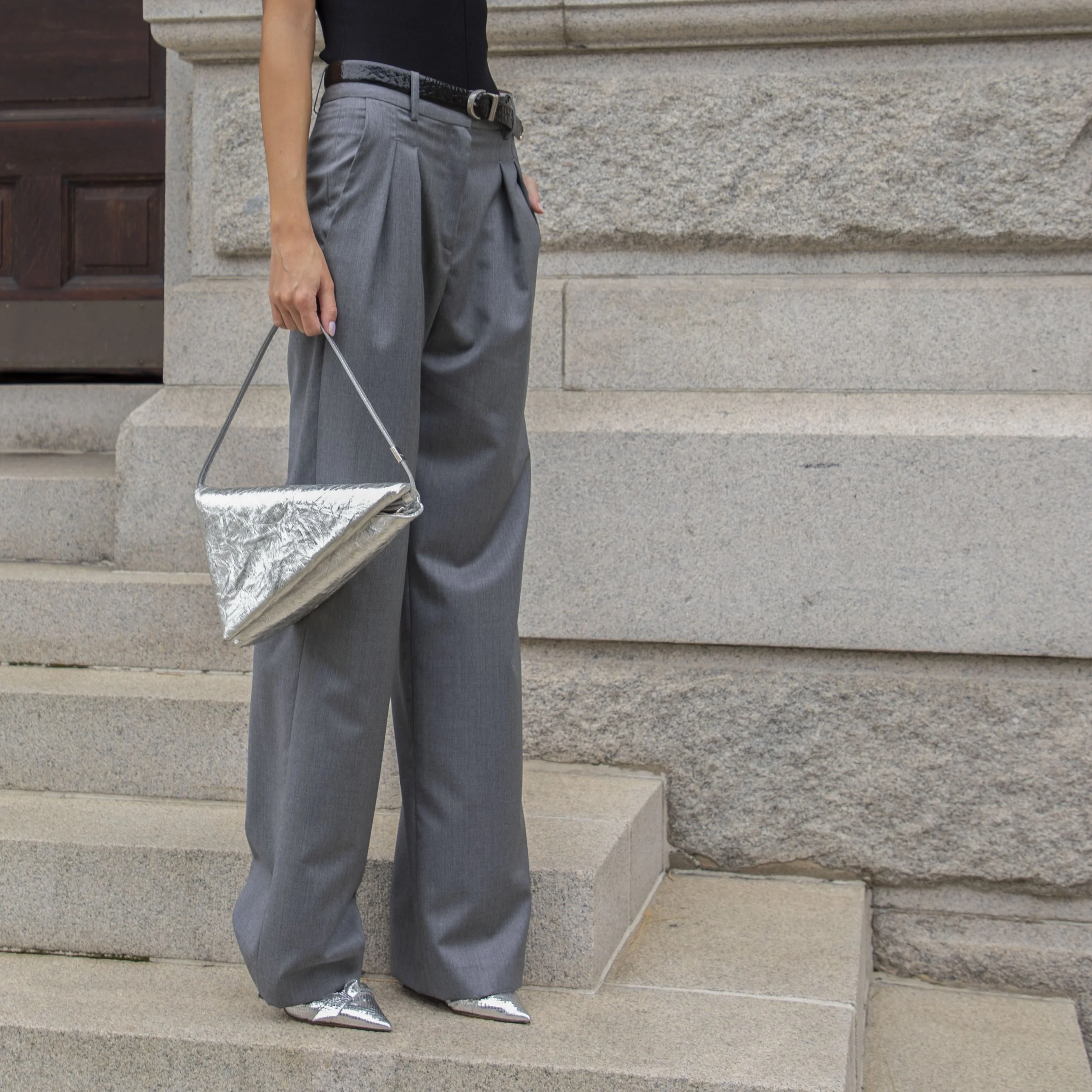 Person standing on stone steps, wearing gray wide-leg pants, a black top, shiny silver pointed-toe shoes, and holding a silver metallic handbag.