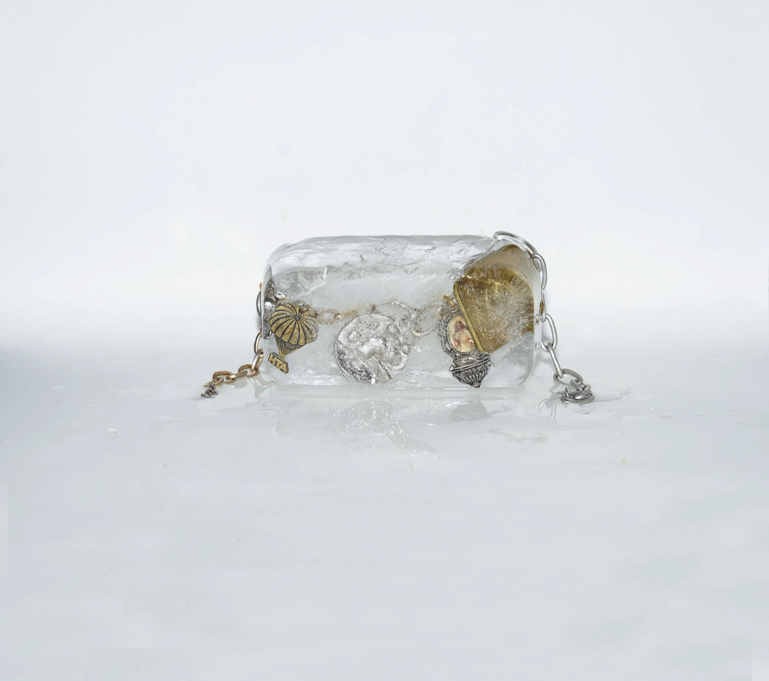 A small handbag made of clear ice with embedded jewelry and embellishments, on a white surface with water droplets.