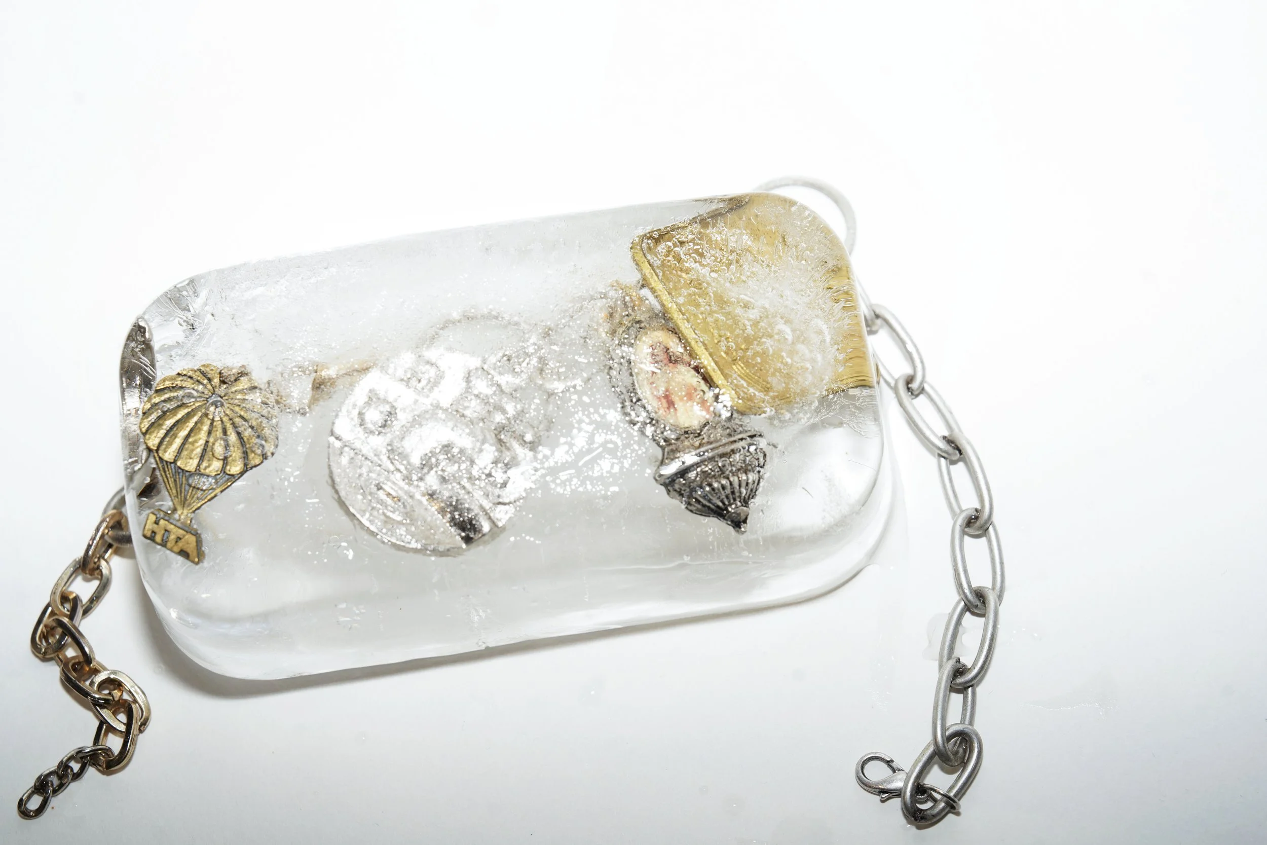 A clear rectangular ice block with embedded metallic pins, charms, and pins, with a chain attached on one side, set against a white background.