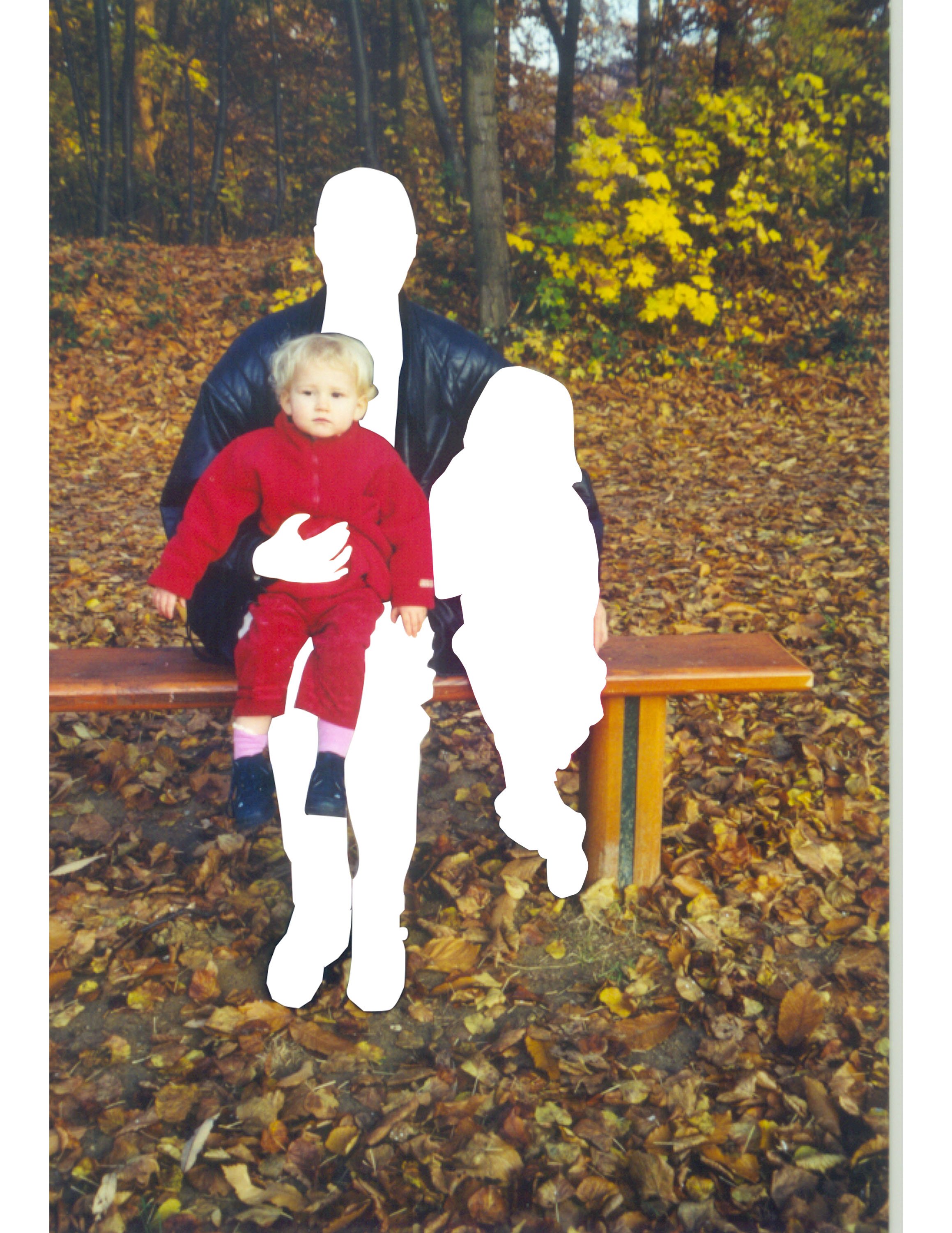 A person in a black leather jacket sitting on a park bench, with two children sitting on their lap. The person and one child's faces are obscured with white, while the child's face is visible. The person is holding a little girl in a red outfit, and there's another child with face obscured sitting next to them. The background shows autumn trees with colorful leaves and fallen leaves on the ground.