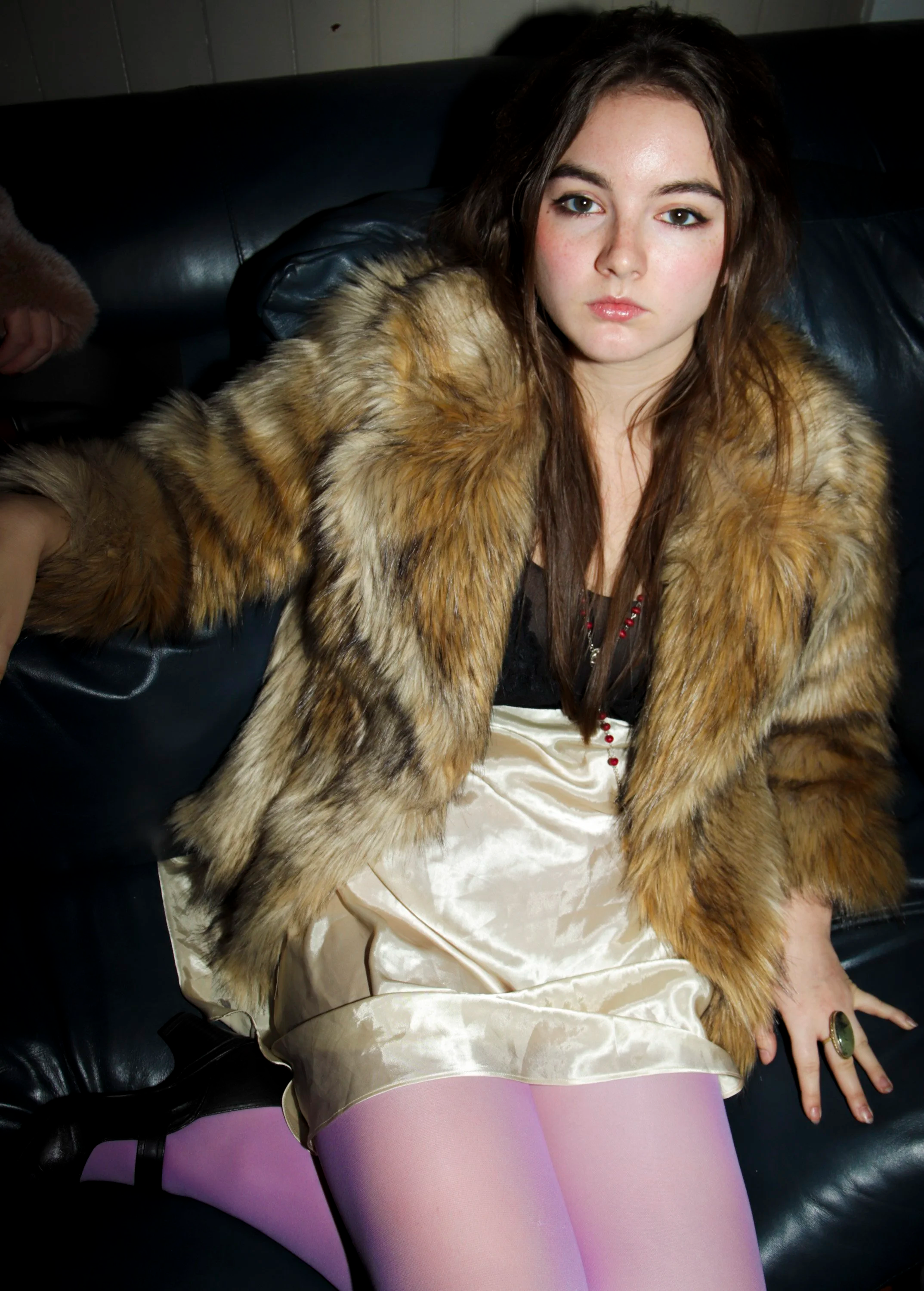 A young woman with long brown hair wearing a fur coat and pink tights sitting on a black leather couch.