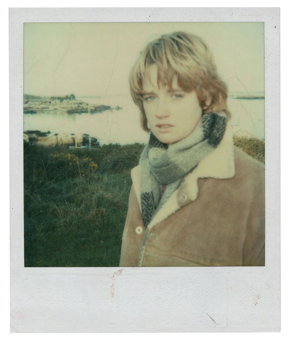 A woman with short blonde hair wearing a tan coat and a gray scarf standing outdoors with a body of water and greenery in the background.