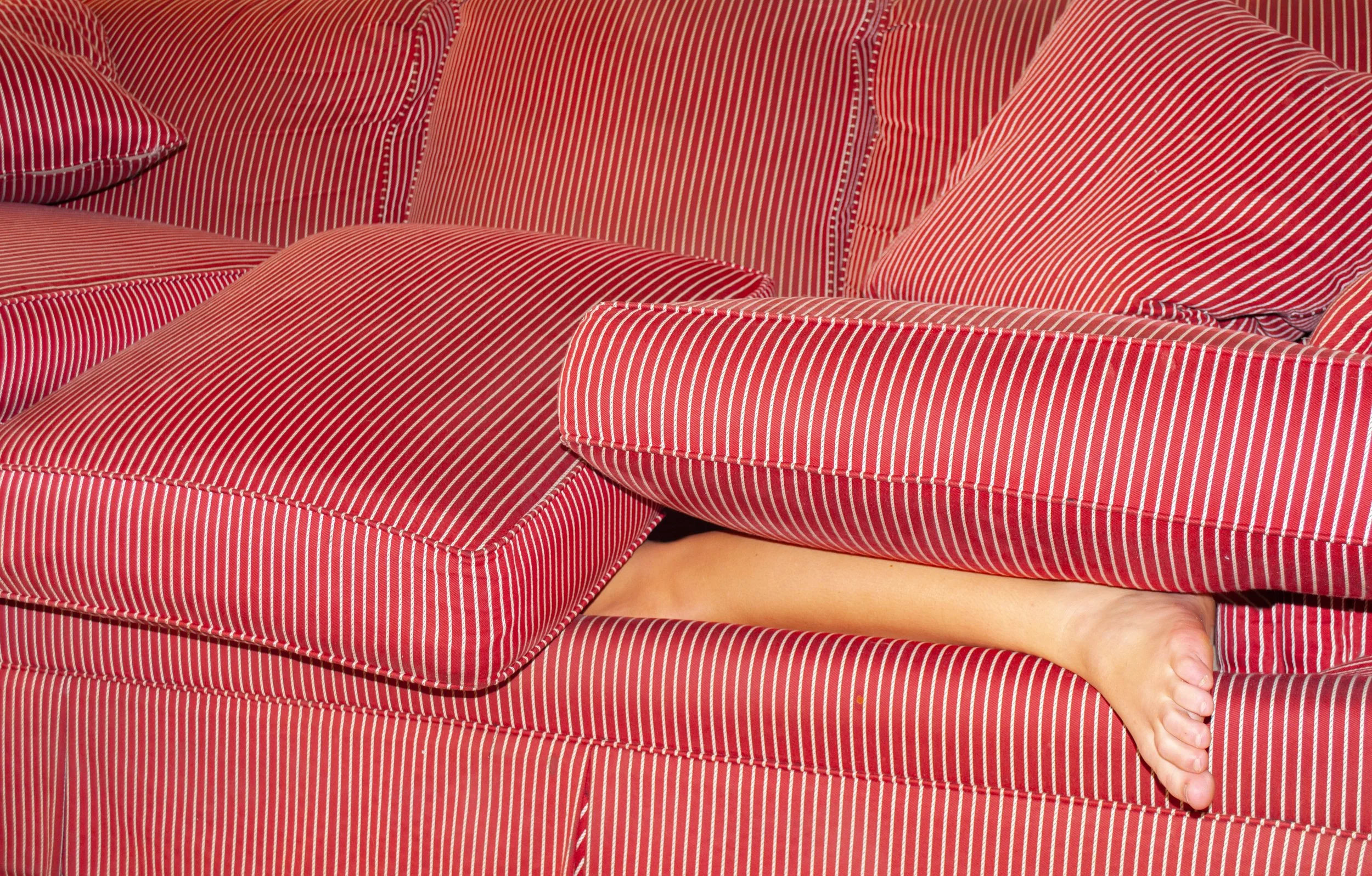 A person lying underneath a red and white striped sofa with only their feet and lower legs visible.