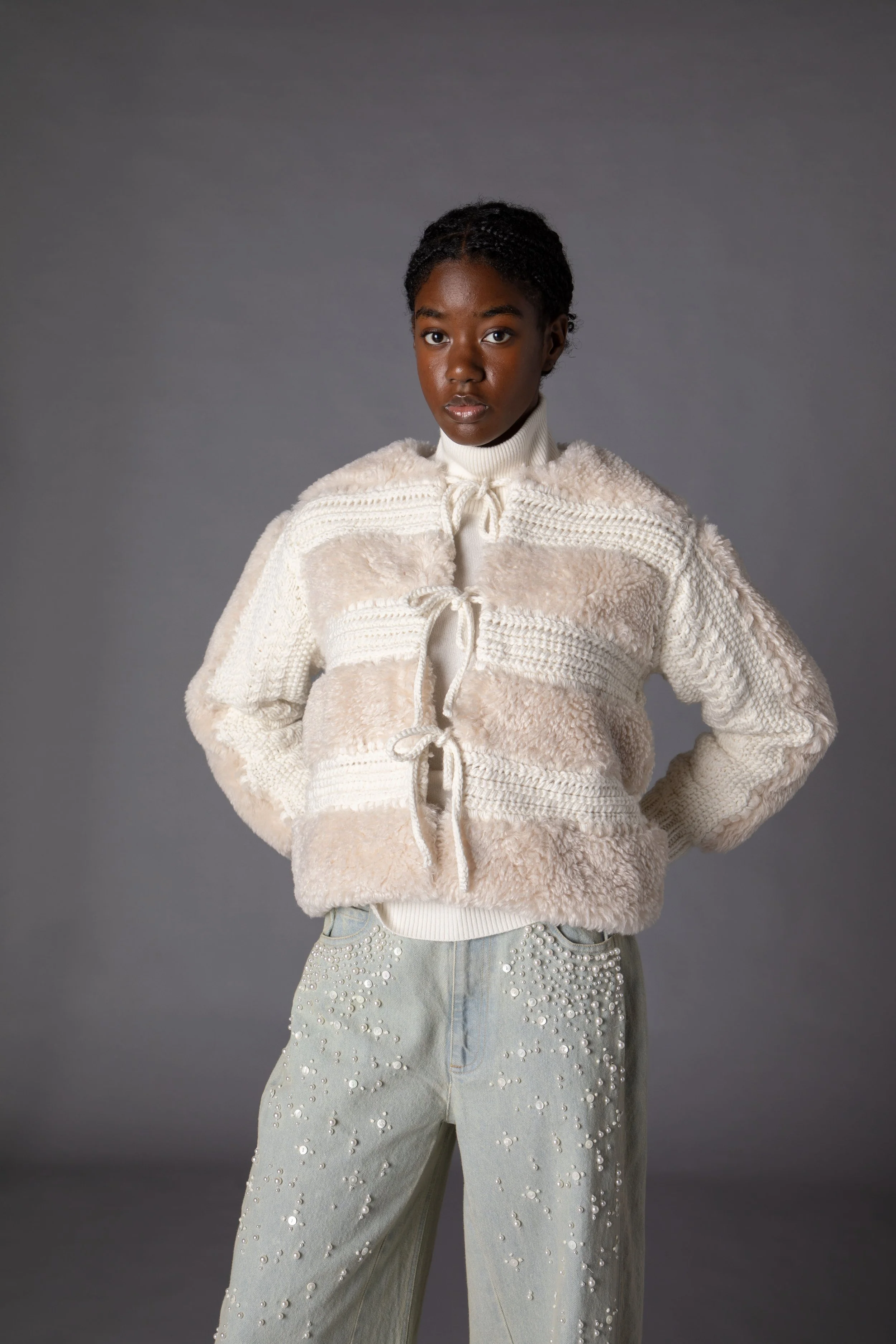 A woman wearing a cozy, cream-colored, patchwork fleece jacket and light-colored jeans decorated with pearls, standing against a plain gray background.