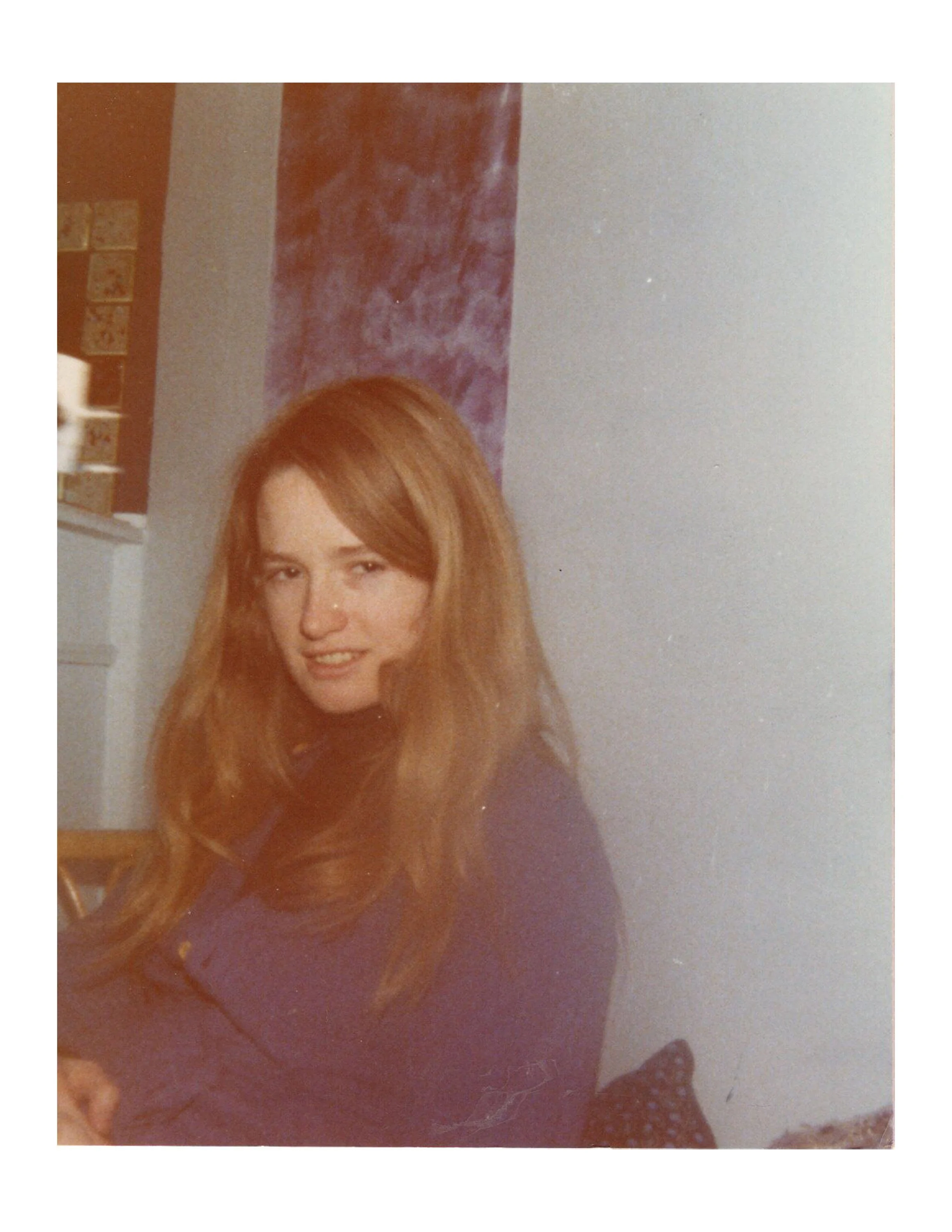 A young woman with long red hair, wearing a dark purple shirt, sitting indoors against a wall with a purple decorative banner and some small framed pictures in the background.