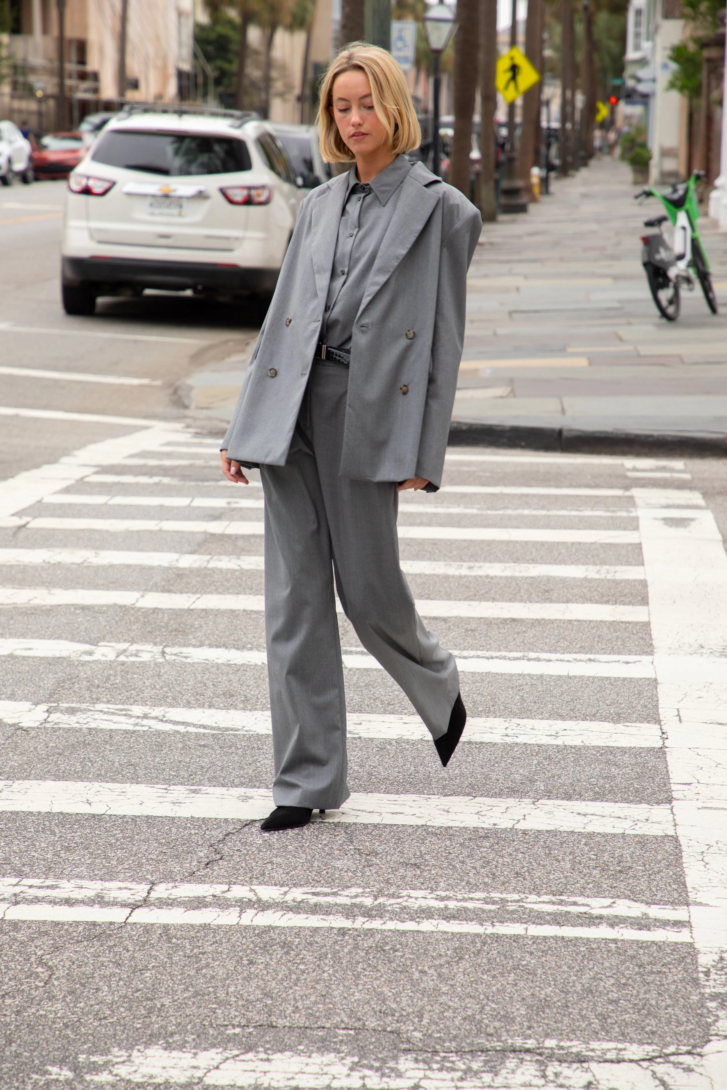 A woman with blonde hair walking across a crosswalk in an urban street, dressed in a gray suit and black shoes.