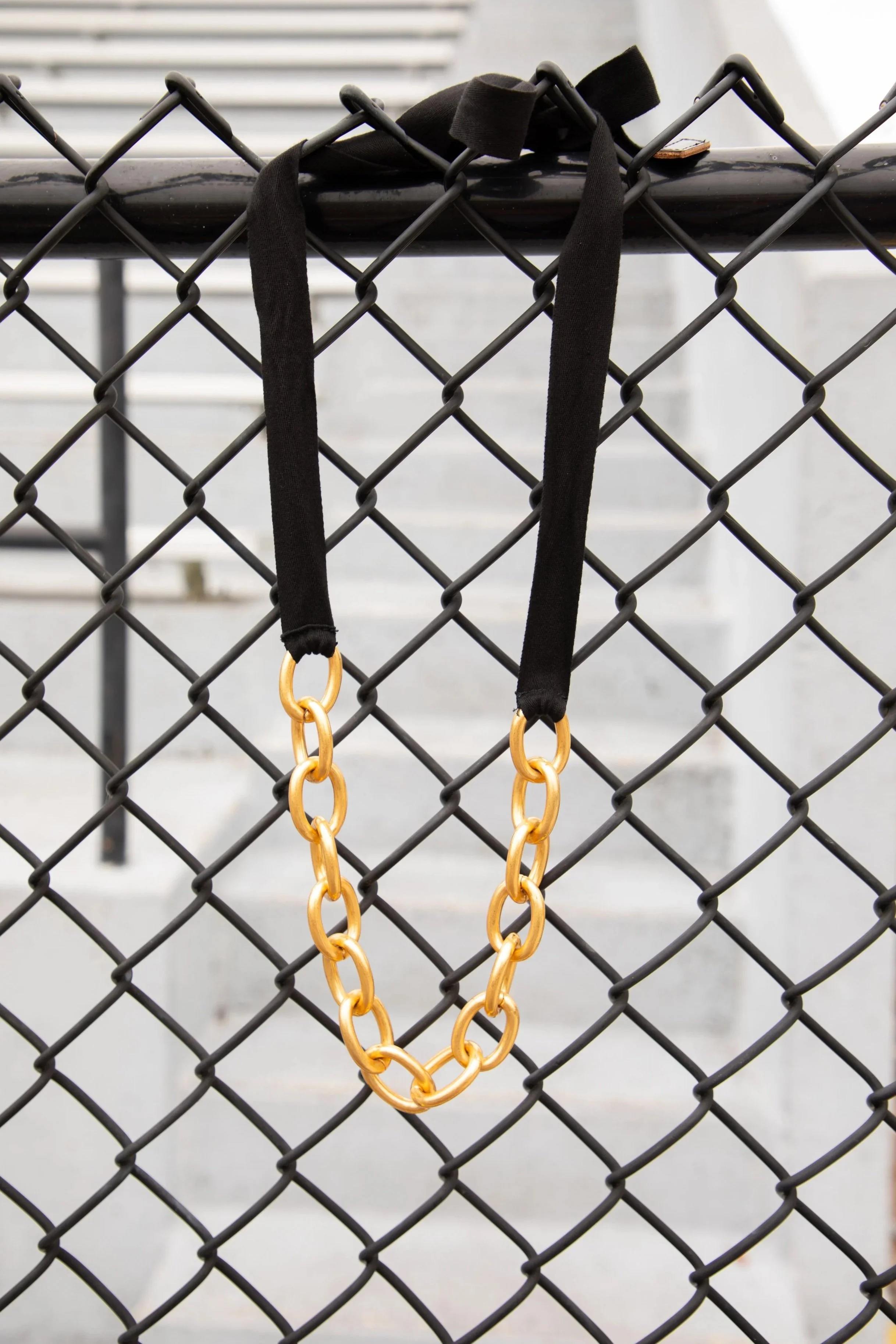 A black fabric necklace with large gold links hanging on a black metal fence.