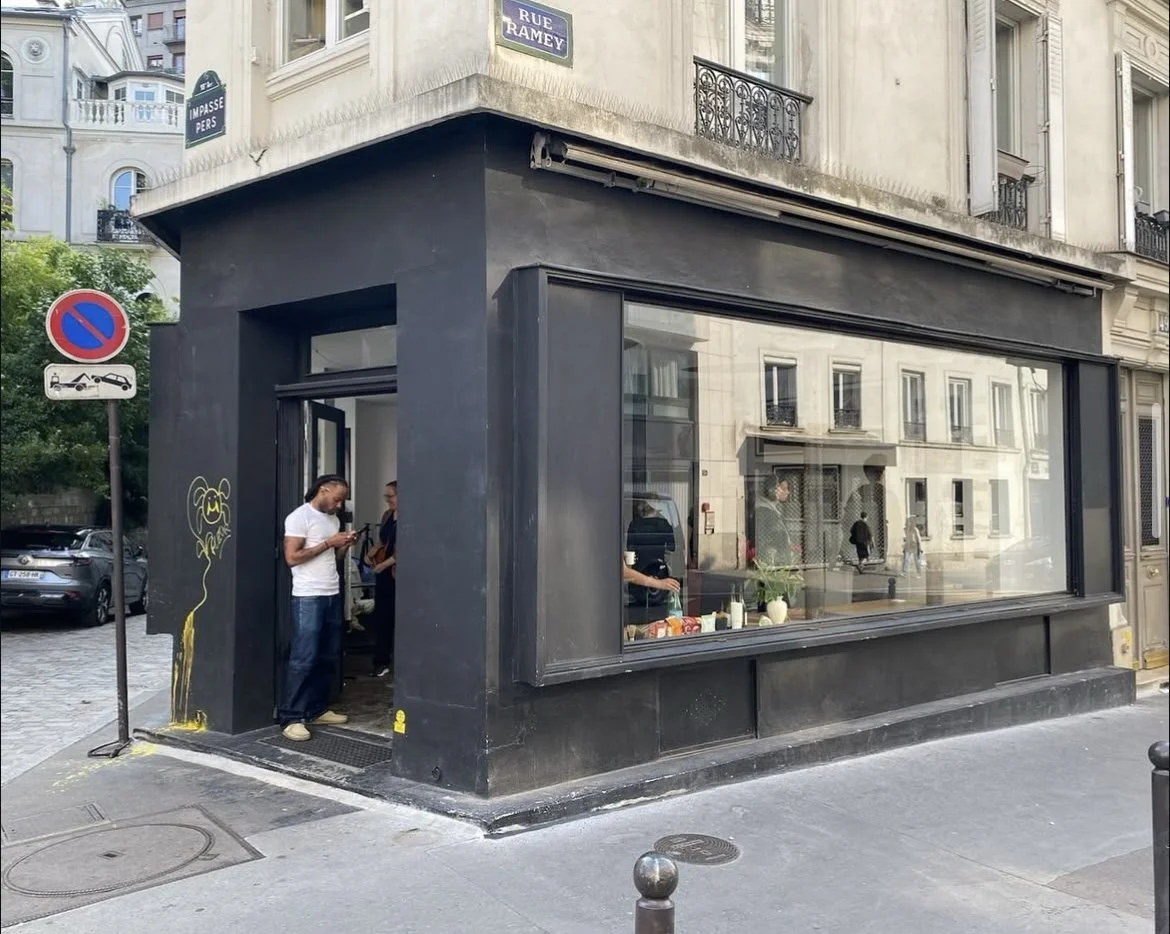 A corner of a building with a large black storefront window and a door, with a man in a white shirt looking at his phone outside. There are street signs, one indicating no parking and another showing a parking restriction, and graffiti on the black w