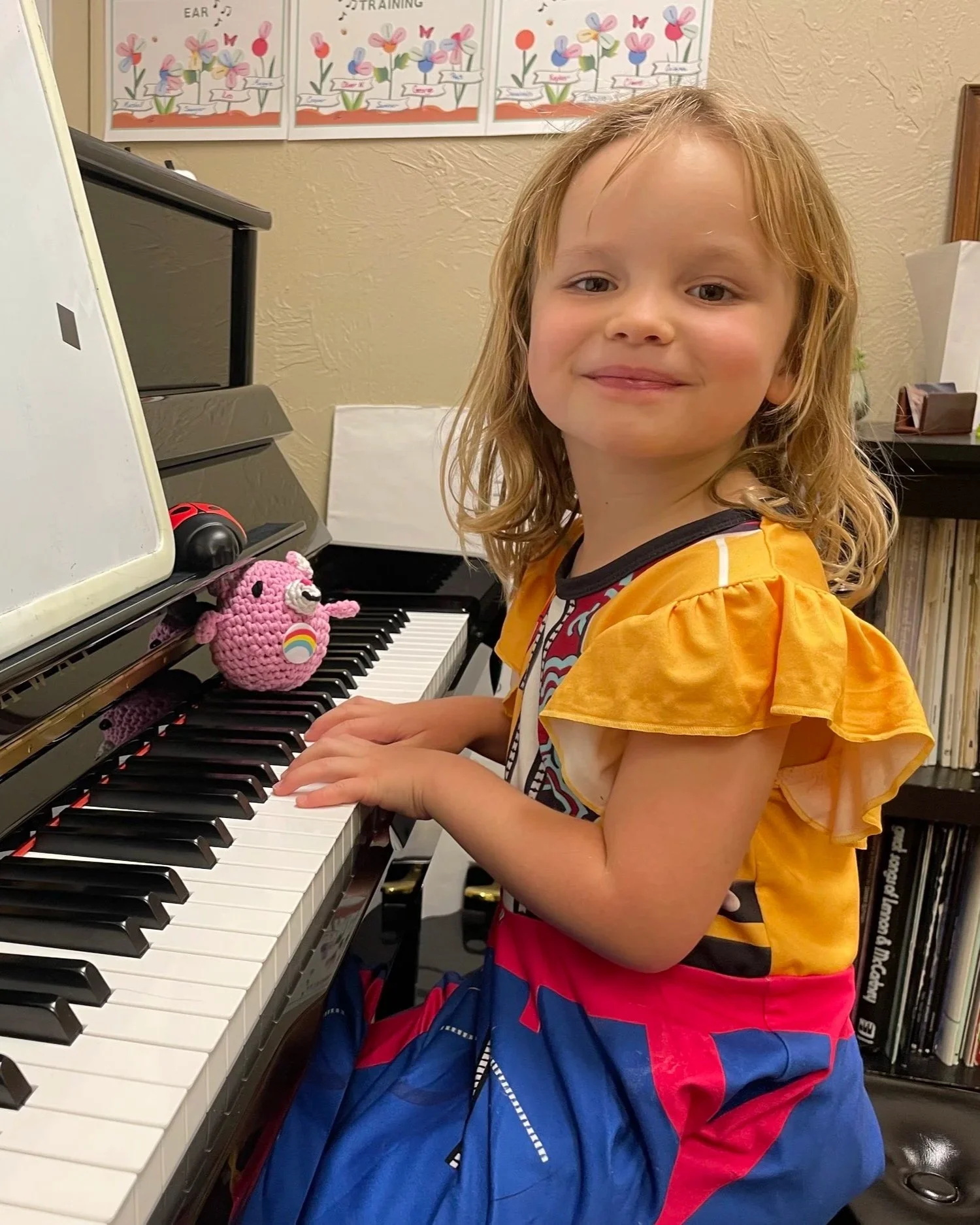 A young girl with shoulder-length blonde hair and a yellow and patterned top sitting at a piano, looking at the camera with a slight smile. There is a pink crocheted hedgehog plush toy on the piano keys.