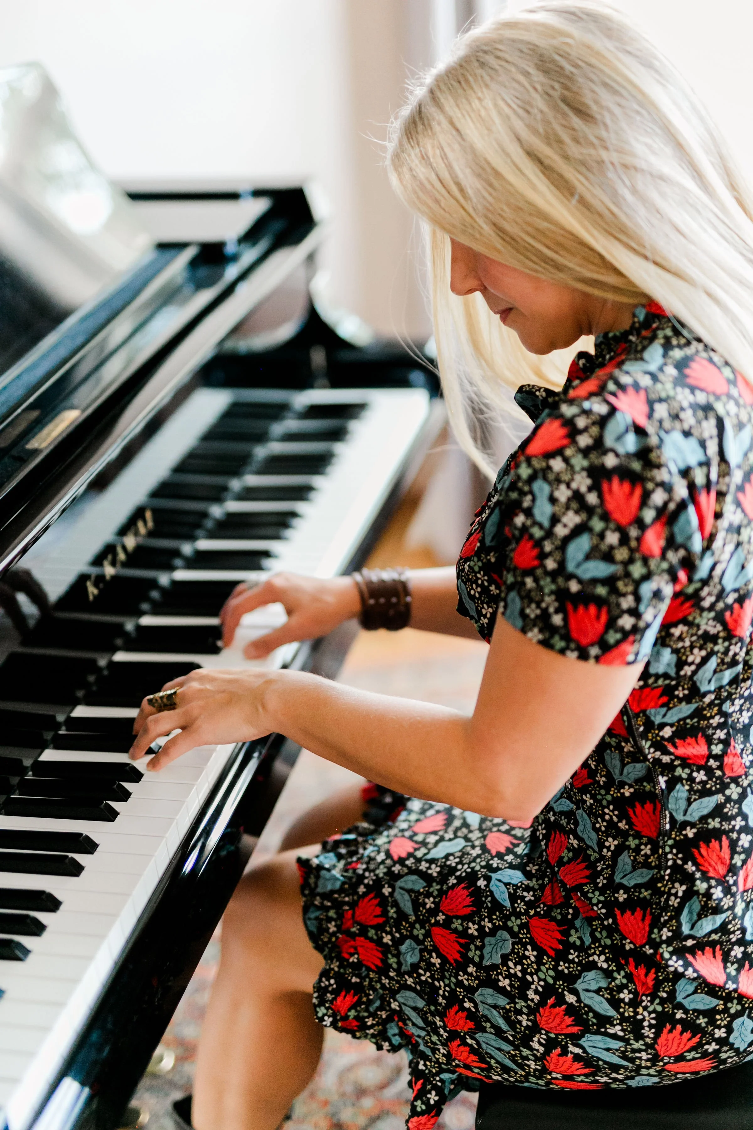A woman with blonde hair wearing a floral dress is playing a black grand piano in a bright room.