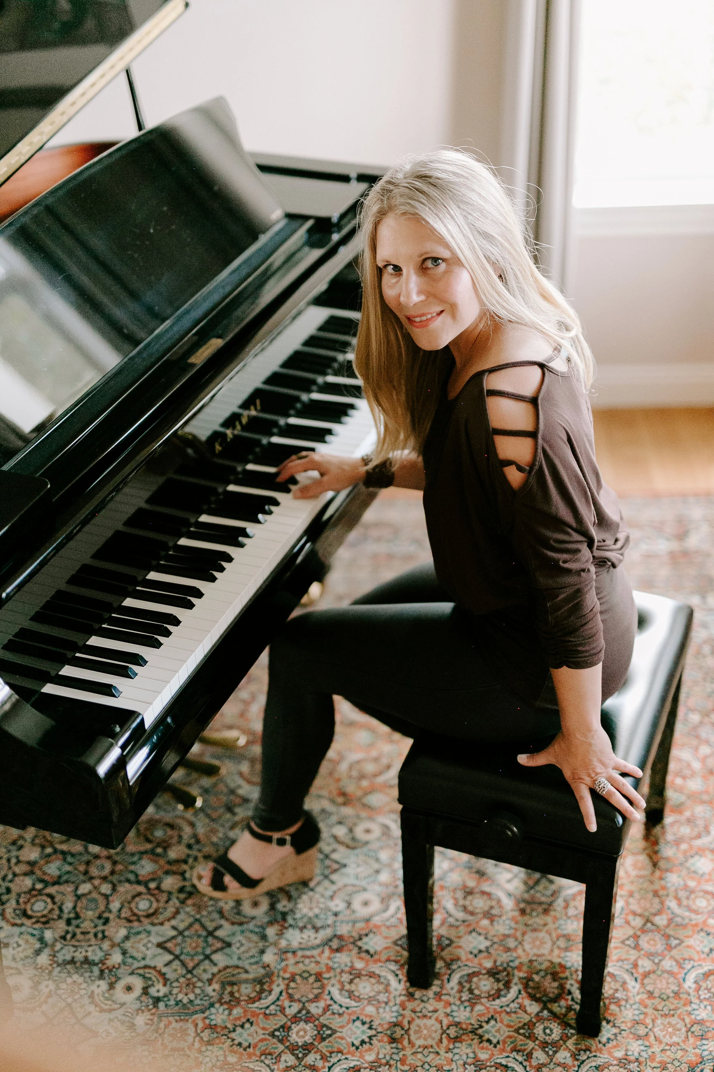 A woman with blonde hair wearing a black top and black pants, sitting on a piano bench, looking up at the camera while playing a black grand piano in a well-lit room.