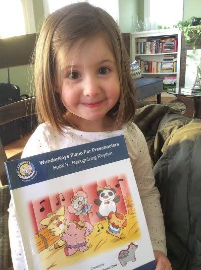 A young girl with brown hair holding a book titled 'WunderKeys Piano for Preschoolers Book 3 - Recognizing Rhythm,' smiling indoors.