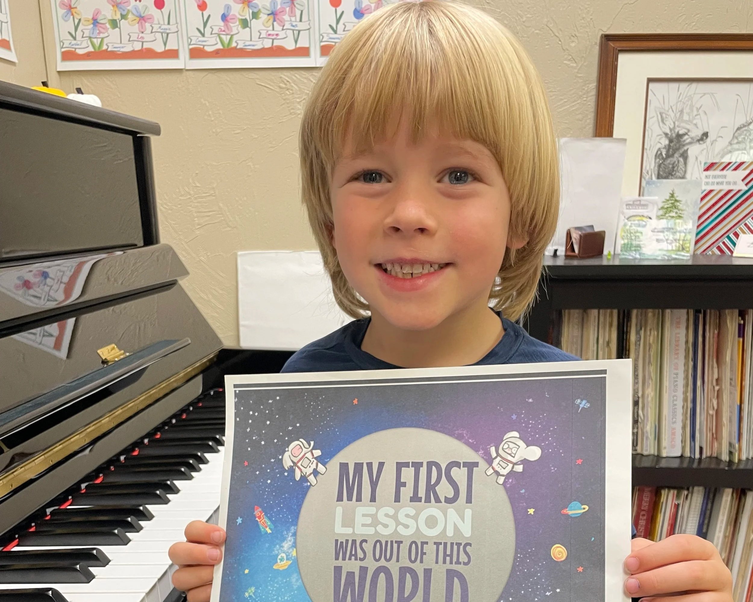 A young boy with blond hair smiling and holding a colorful space-themed sign that reads, "My first lesson was out of this world," standing in front of a piano and a bookshelf in a room decorated with children's artwork.