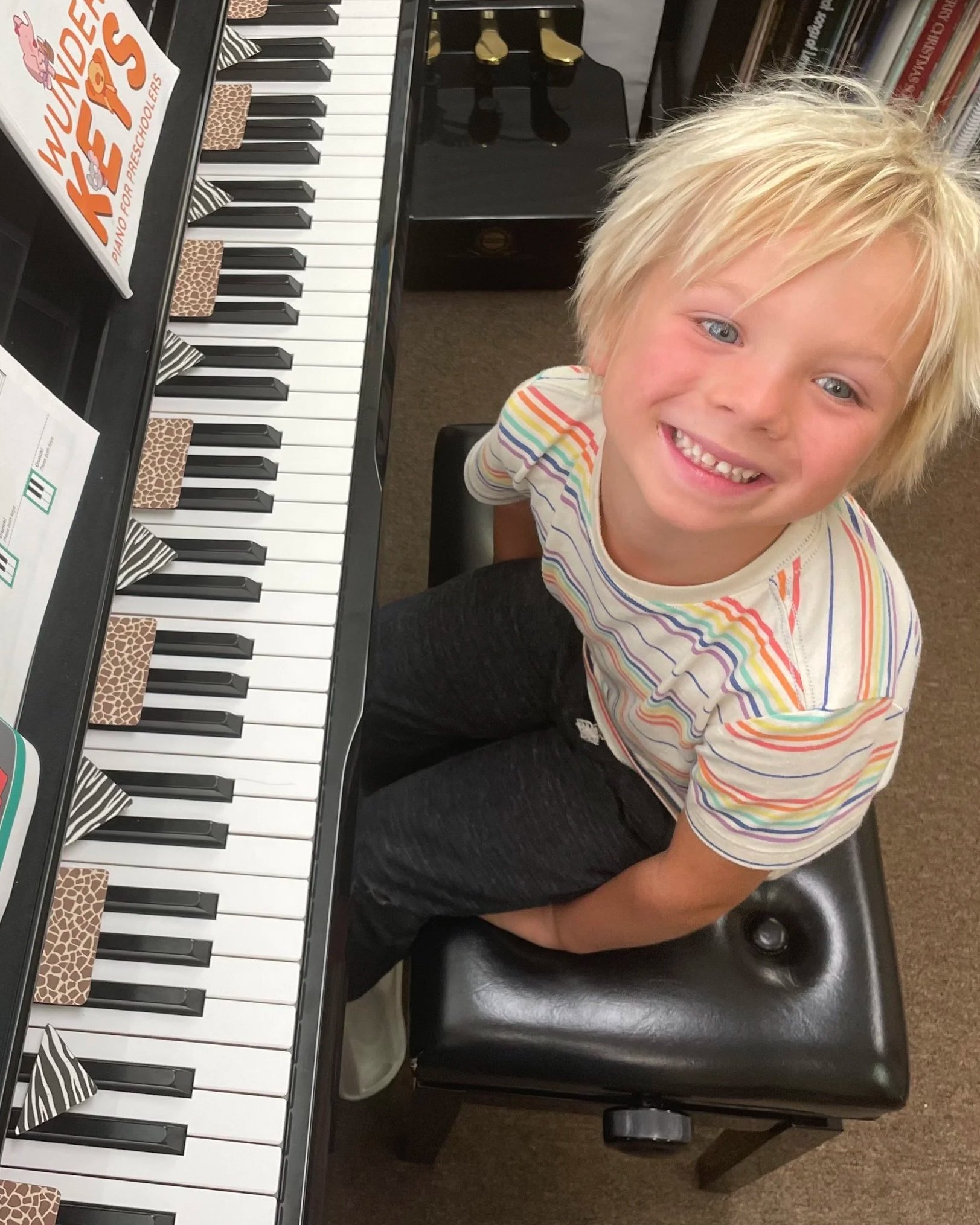 A young boy with blonde hair and a rainbow-striped shirt, sitting at a black upright piano, looking up and smiling at the camera.
