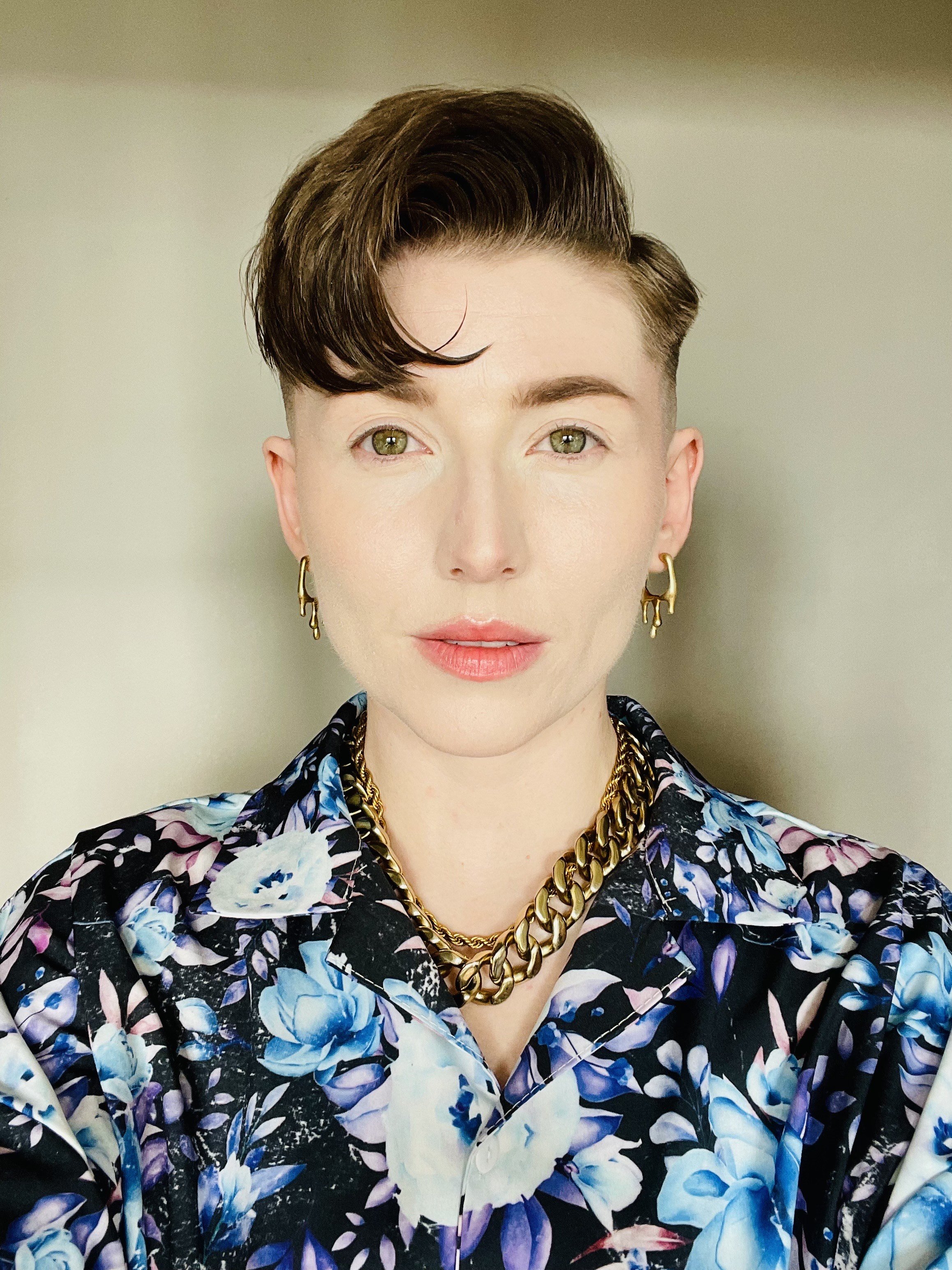 Portrait of a person with short, dark, styled hair, wearing gold earrings, a chunky gold chain necklace, and a floral patterned shirt with shades of purple, blue, and white.