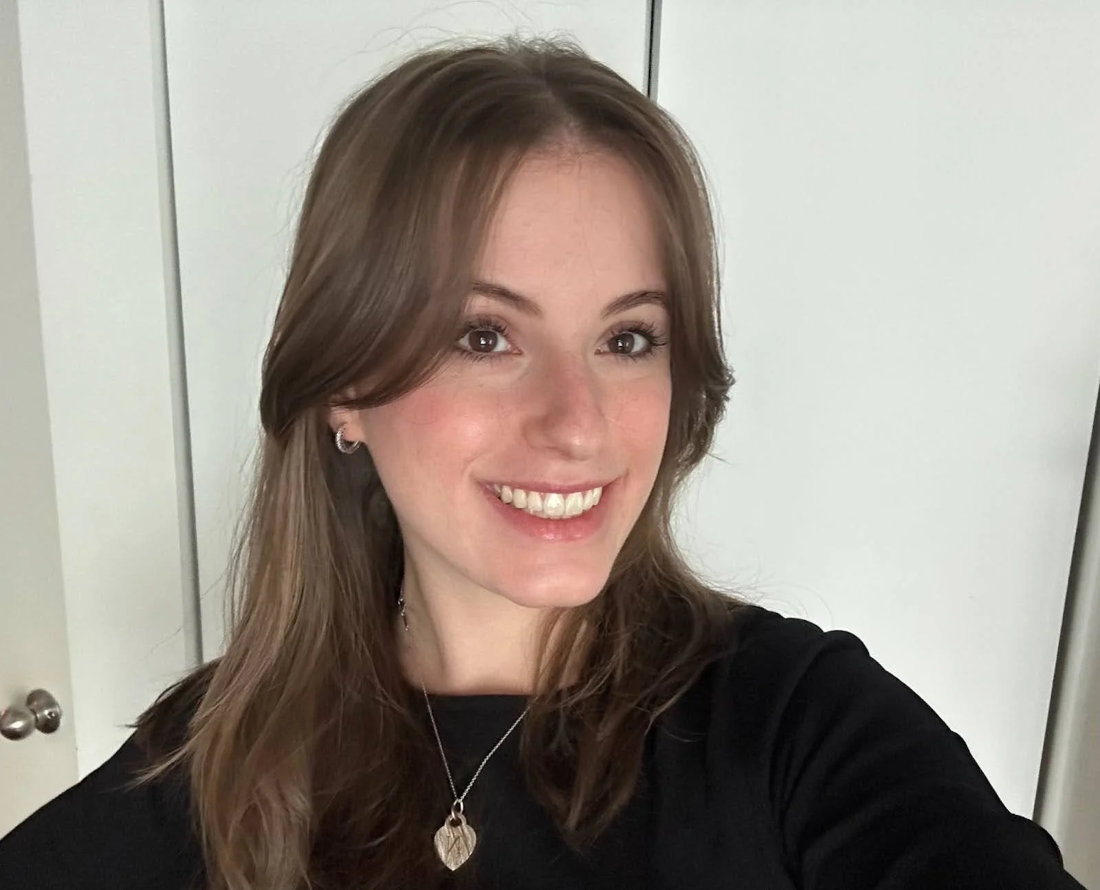 A woman with long brown hair, smiling, wearing a black shirt and jewelry, standing in front of a white door.