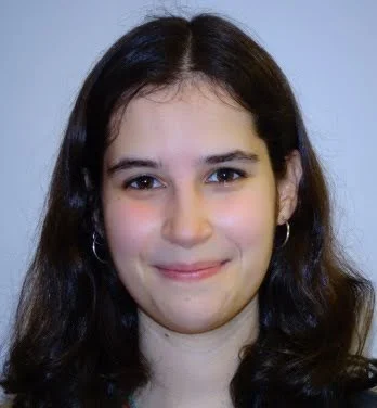 Close-up of a young woman with dark hair, smiling, wearing earrings, against a plain background.