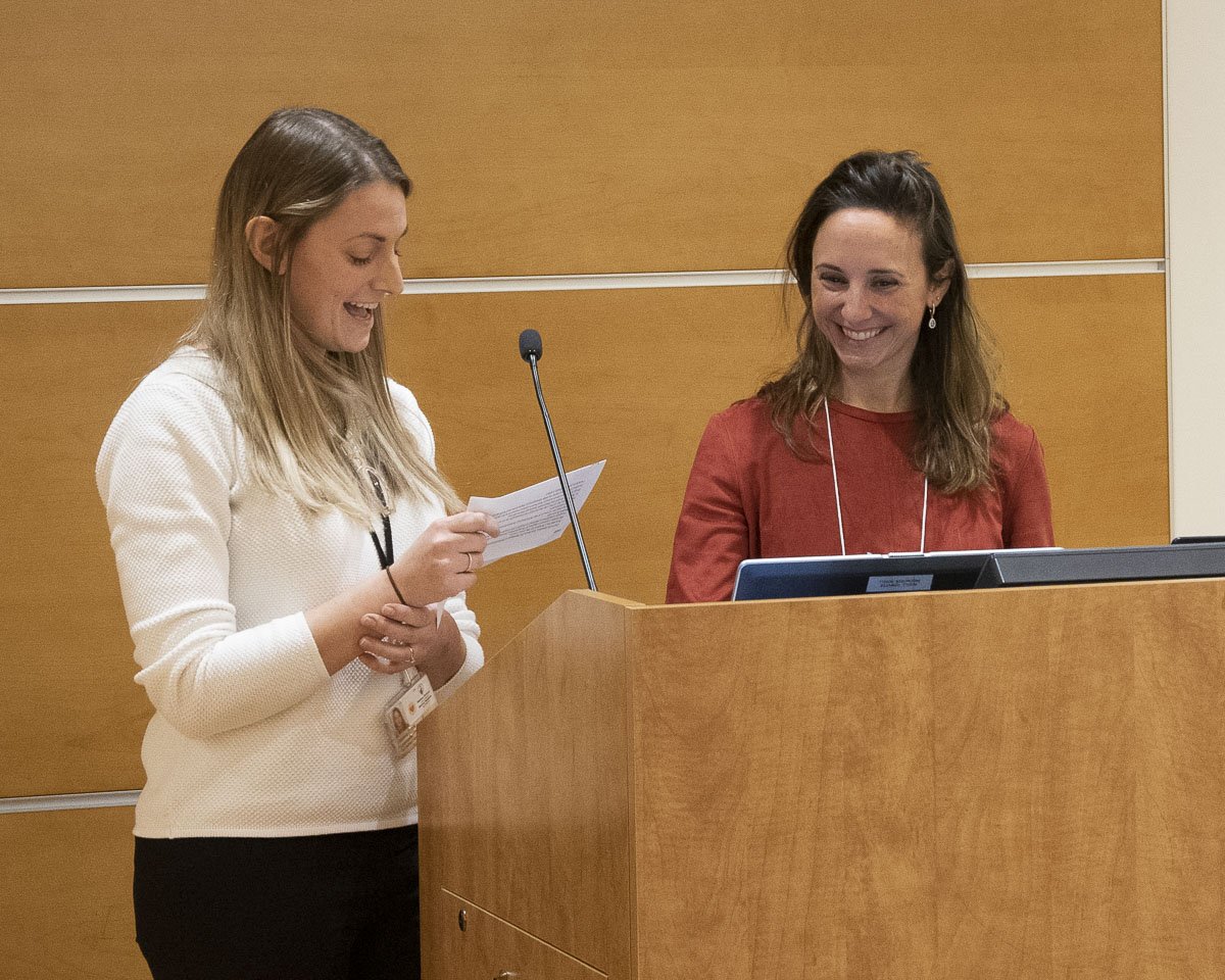 Two women smiling and talking at a podium in a conference room.