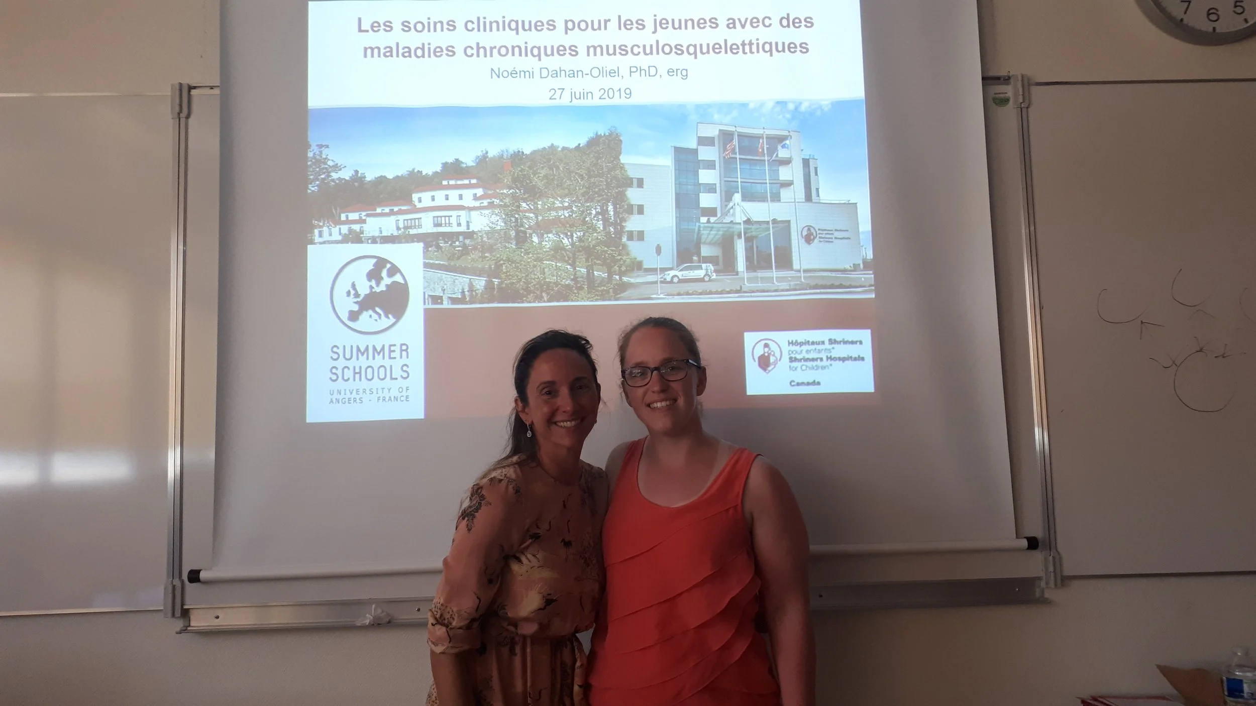Two women smiling in front of a projection screen in a classroom setting. The screen displays a presentation titled in French about clinical care for young people with musculoskeletal chronic diseases, with logos from Summer Schools University of Ang