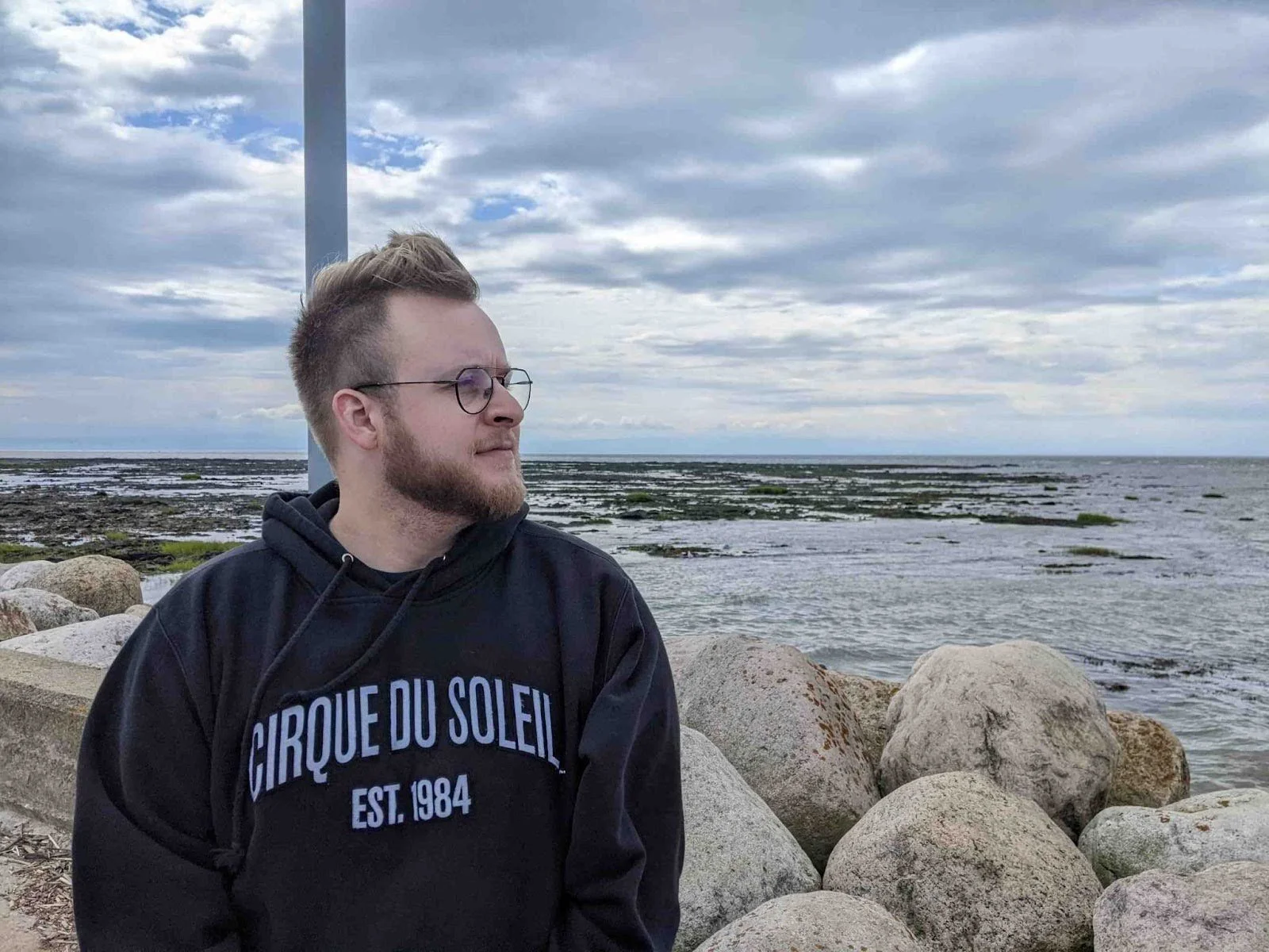 A young man with glasses and a beard, wearing a black hoodie with 'CIRQUE DU SOLEIL EST. 1984' written on it, standing by rocks along a shoreline, looking to the side with a cloudy sky in the background.