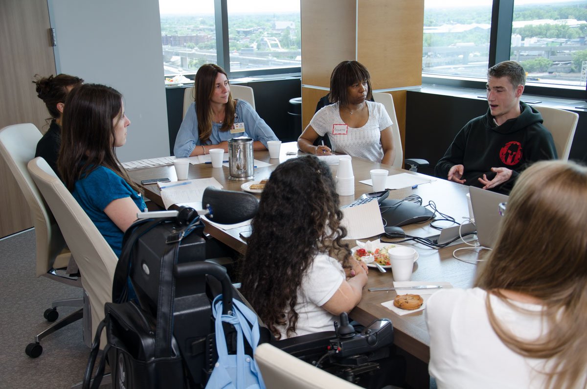 Group of diverse people sitting around a conference table, participating in a meeting, with snacks and drinks, in a high-rise office with large windows showing an urban view.