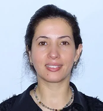 A woman with dark hair pulled back, wearing a black top with a beaded necklace, smiling against a light blue background.