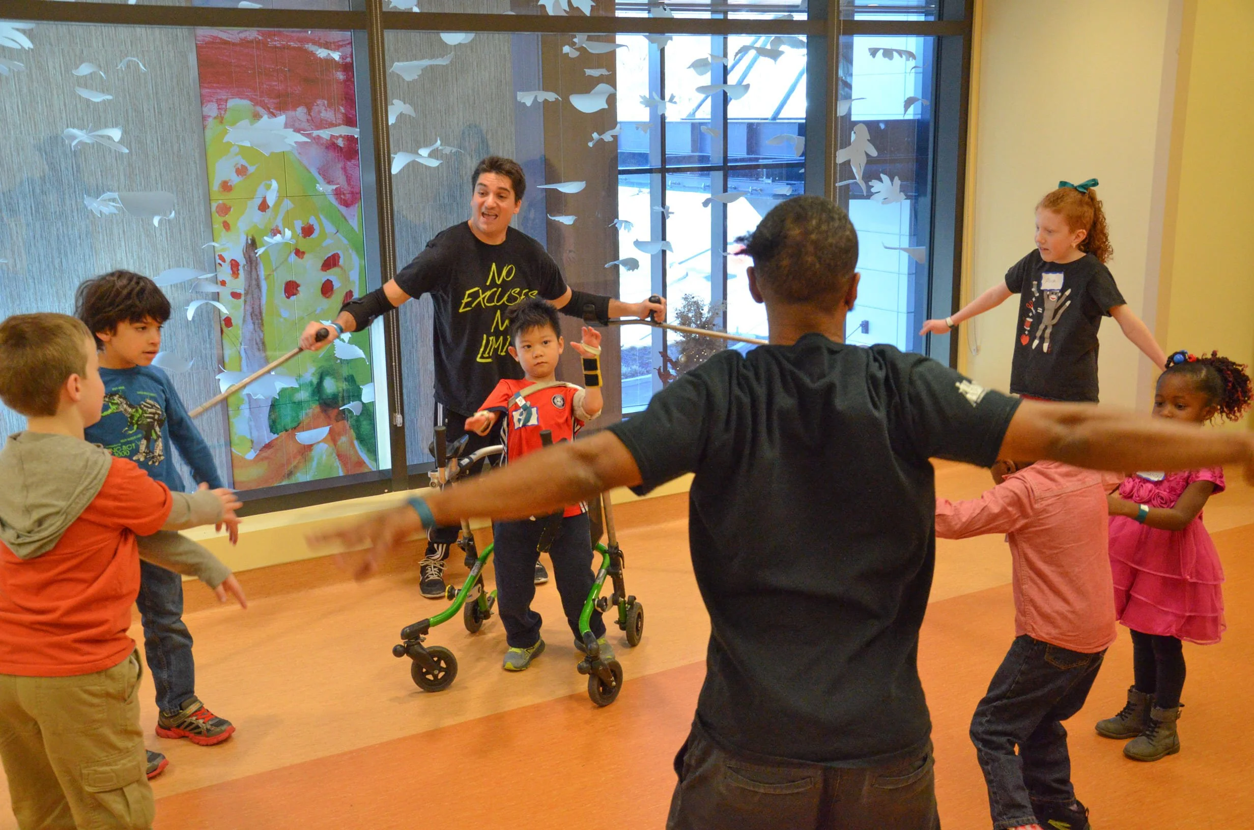 Children and adults participating in a group activity indoors, holding hands and forming a circle, with a person leading the activity, in front of large windows with decorations.