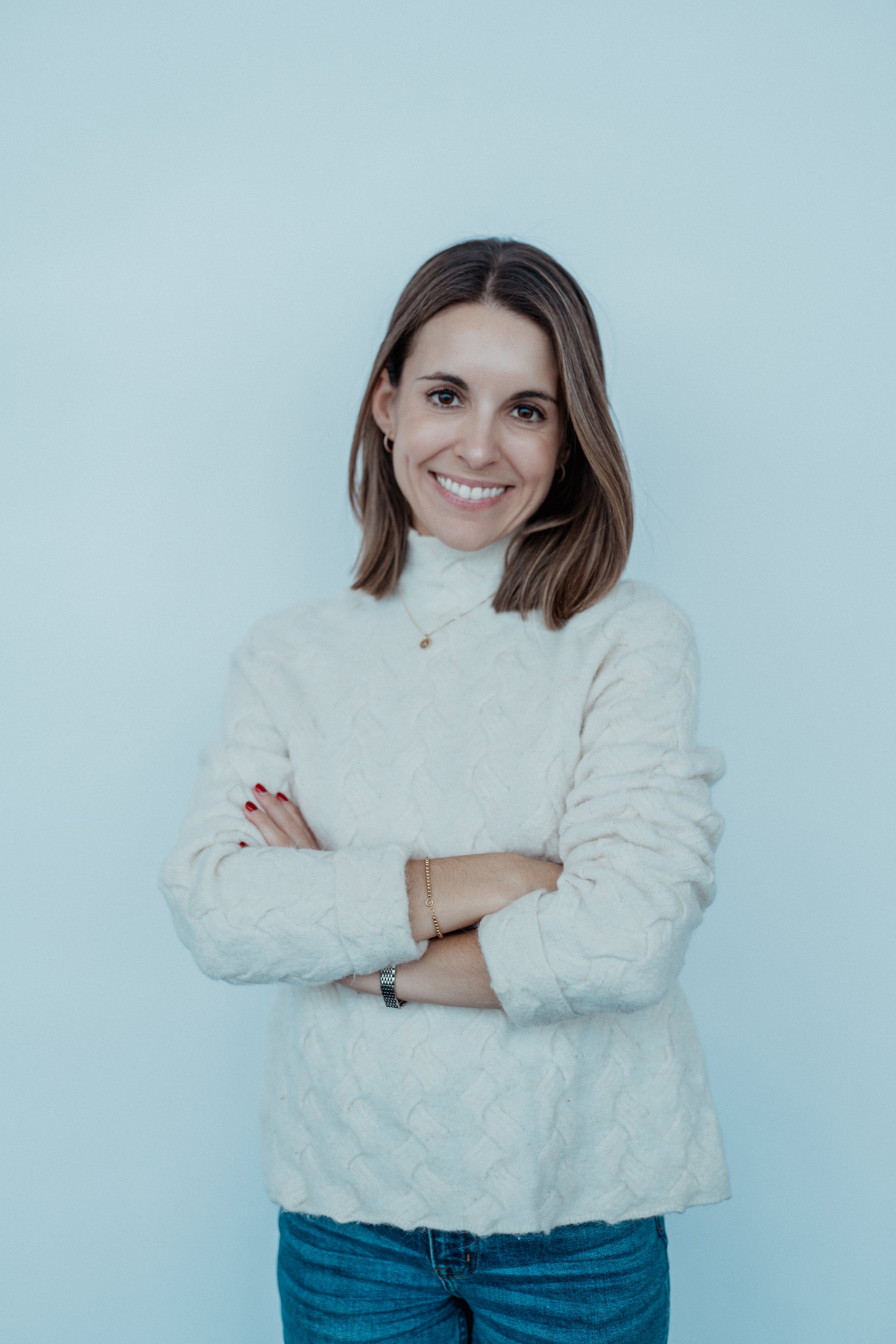 A smiling woman with brown hair, wearing a white textured sweater and blue jeans, crossing her arms while standing against a light blue background.