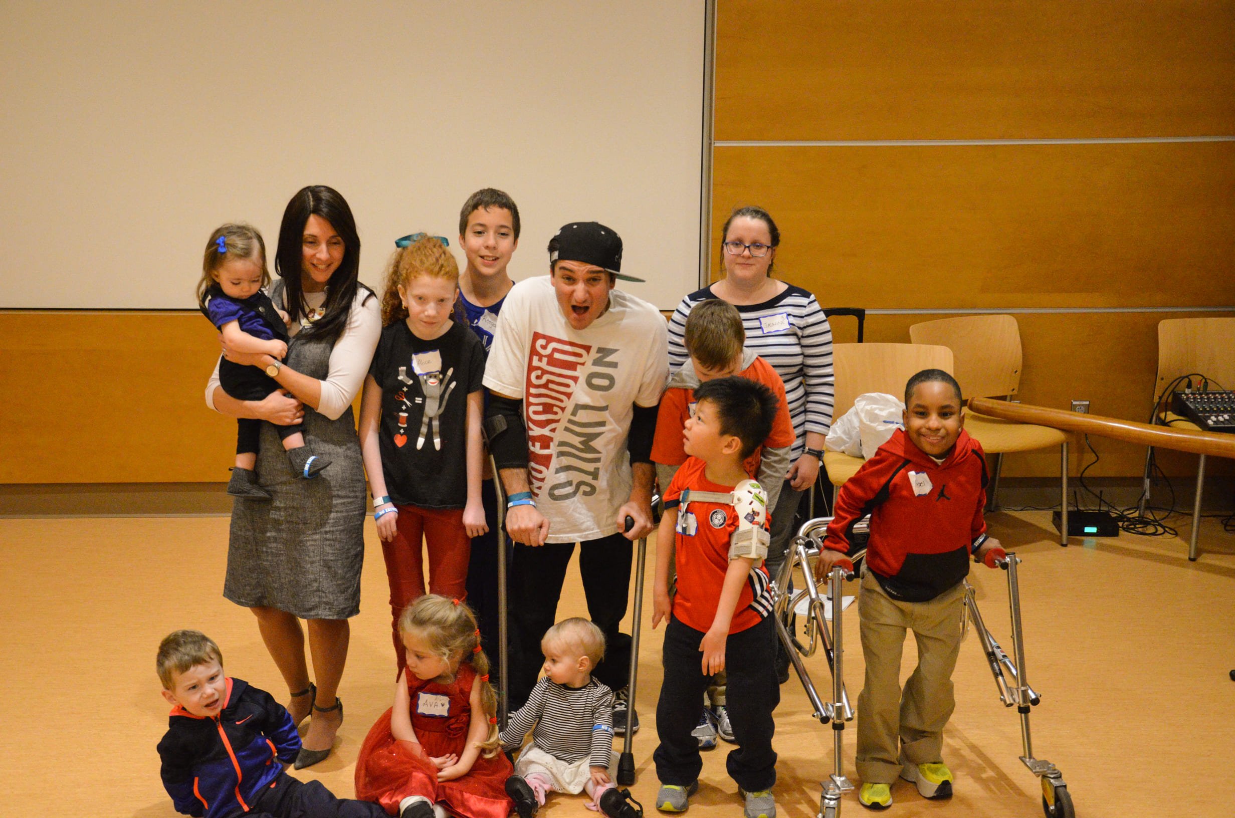 A group of children and two adults, one woman and one man with crutches, posing together in a room with a wooden floor and beige walls, some children on the floor and some standing, smiling at the camera.