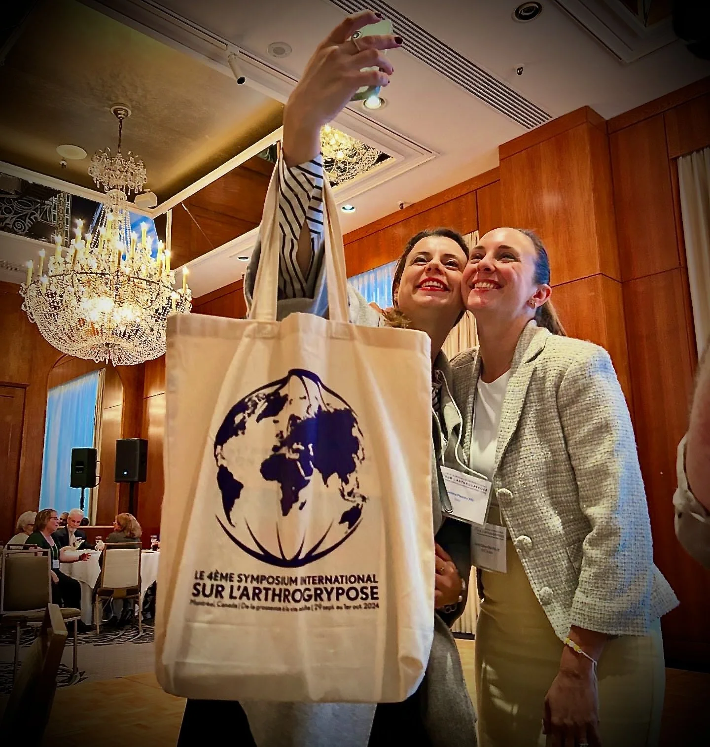 Two women taking a selfie at a conference, one holding a tote bag with a logo and text about an international symposium on arthrogryposis. The background shows a conference room with chandeliers and seated attendees.