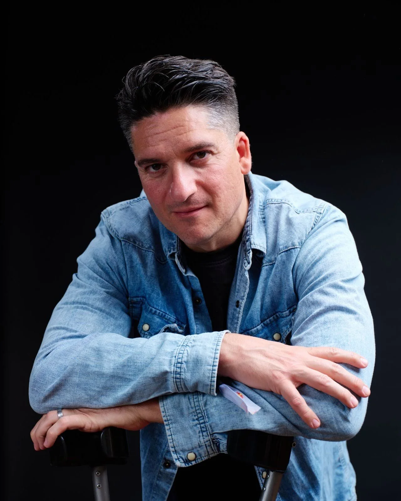 Man with dark hair wearing a denim jacket, sitting with arms crossed on a crutch against a black background.
