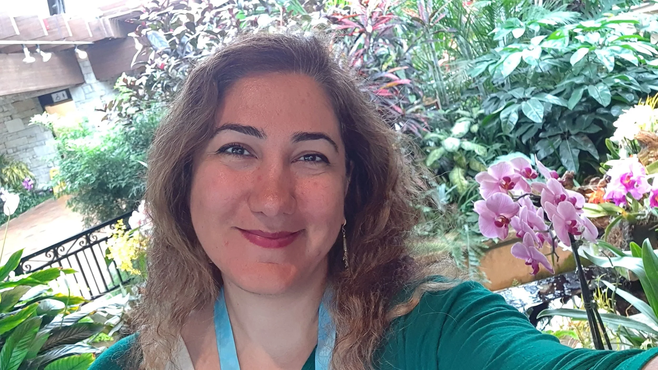 A woman with curly hair smiles at the camera in a lush indoor garden with orchids and various green plants.