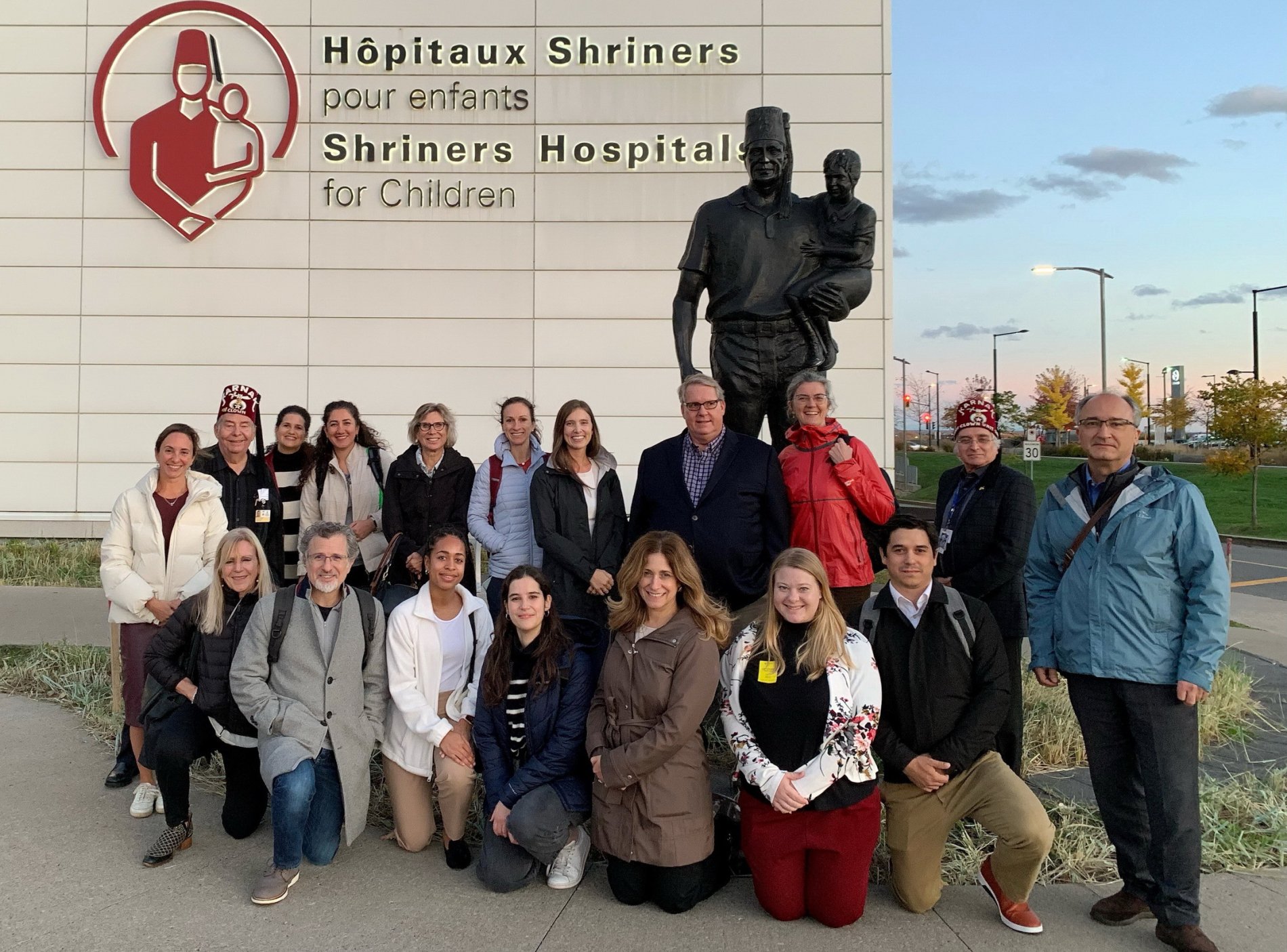 Group of people posing in front of a sign and statue outside Shriner's Hospitals for Children, with a large logo, and a statue of a man dressed as a Shriner in a city street setting during winter.