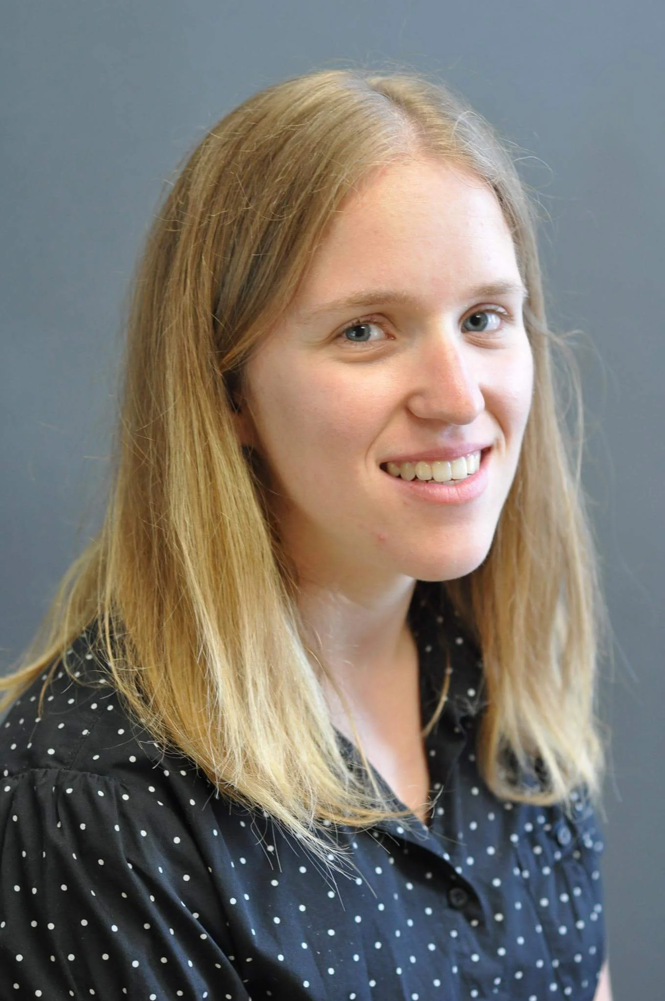 Portrait of a woman with long blond hair, light skin, and blue eyes, wearing a black shirt with white polka dots, standing against a gray background.