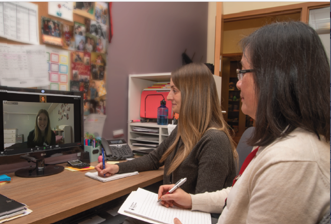 Two women sitting at a desk during a video conference with a third woman on the computer screen, taking notes in a notebook.