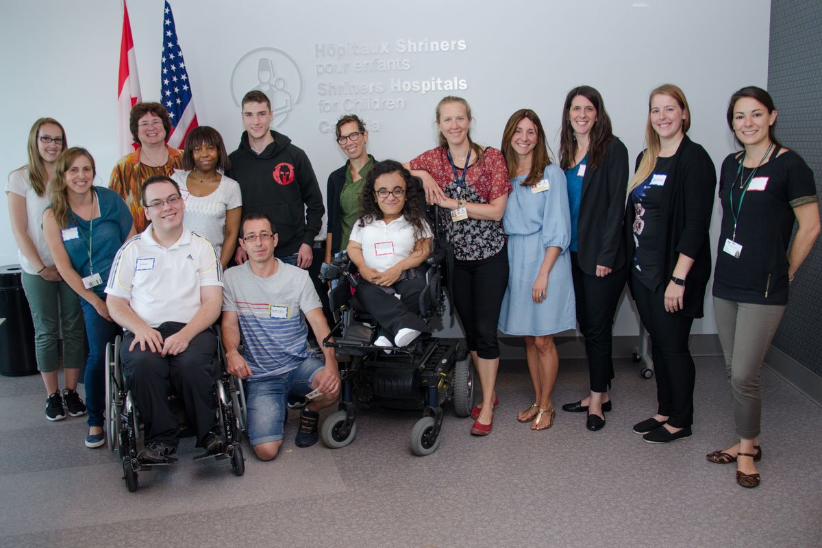 Group photo of diverse young people, some in wheelchairs, at a hospital event with U.S. and Canadian flags in the background.
