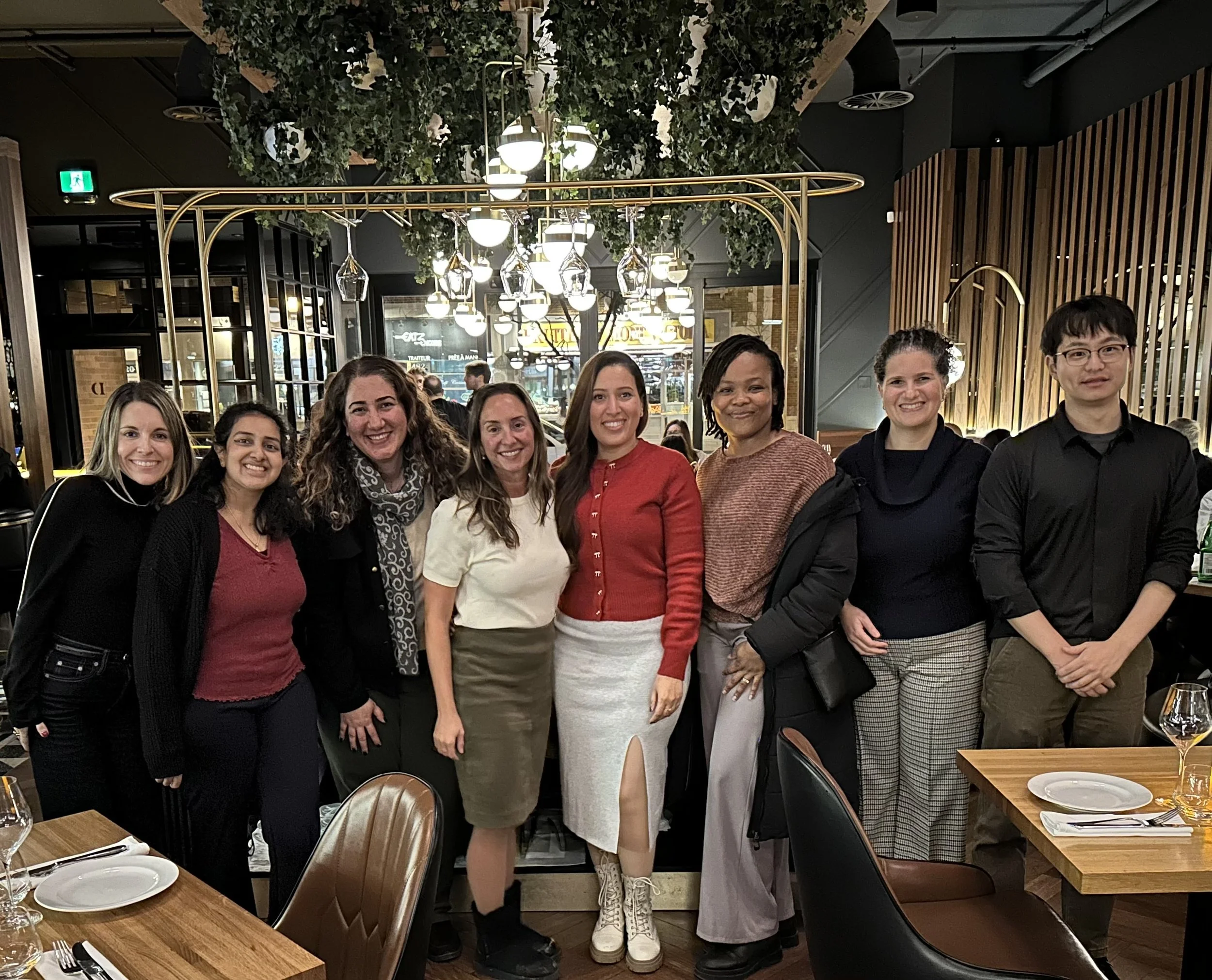A group of nine women and one man standing together in a restaurant, smiling at the camera with tables and wine glasses in the foreground.