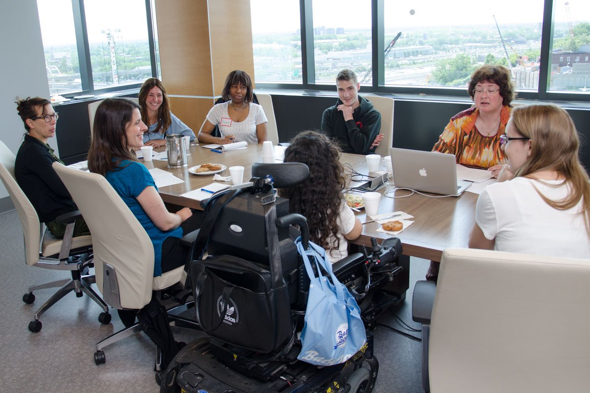 A diverse group of eight women and one person in a wheelchair gathered around a conference table in a modern office with large windows, having a meeting or discussion.