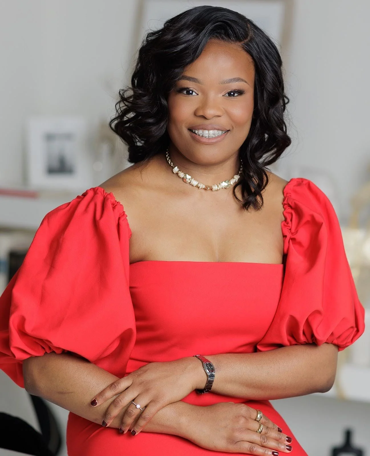 A woman with shoulder-length black hair, wearing a red dress with puffed sleeves, a pearl necklace, a watch, and rings, smiling in an indoor setting.