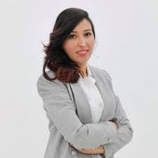 A woman with dark hair, wearing a gray blazer over a white shirt, standing against a plain background. She has her arms crossed and is smiling.